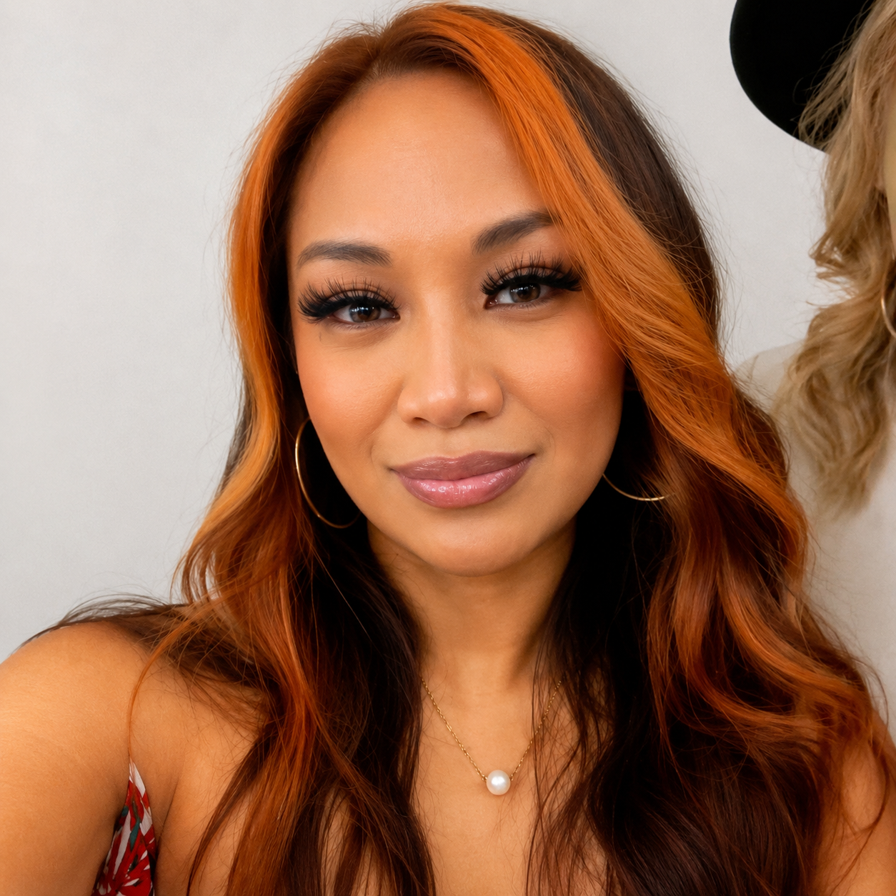 Close-up of a woman with long wavy red and brown hair, wearing hoop earrings, makeup, and a pearl necklace, smiling at the camera.
