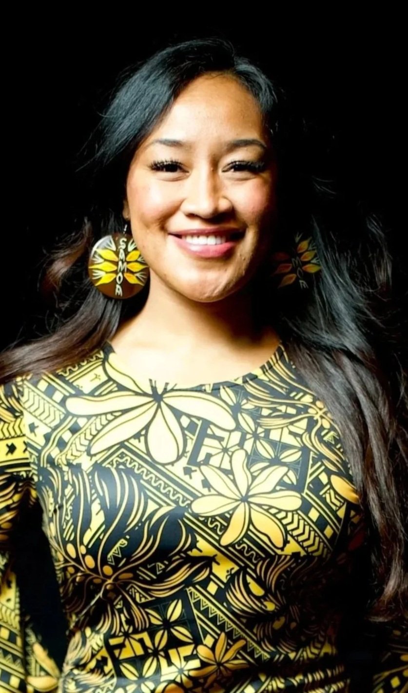 Smiling woman with long black hair, wearing large yellow earrings and a yellow and black patterned dress against a black background.