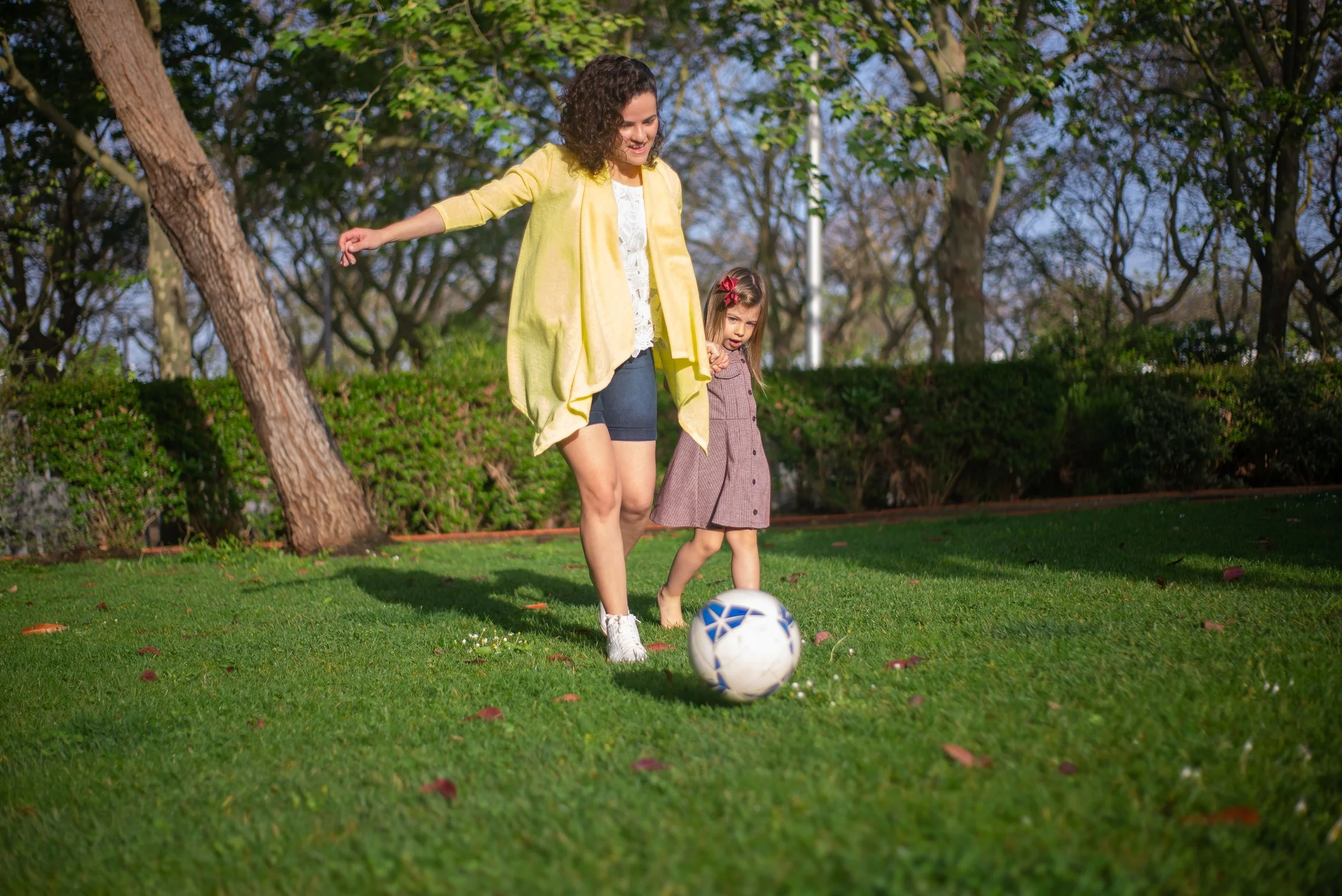 A mother guiding her young daughter as they play soccer together in a park.