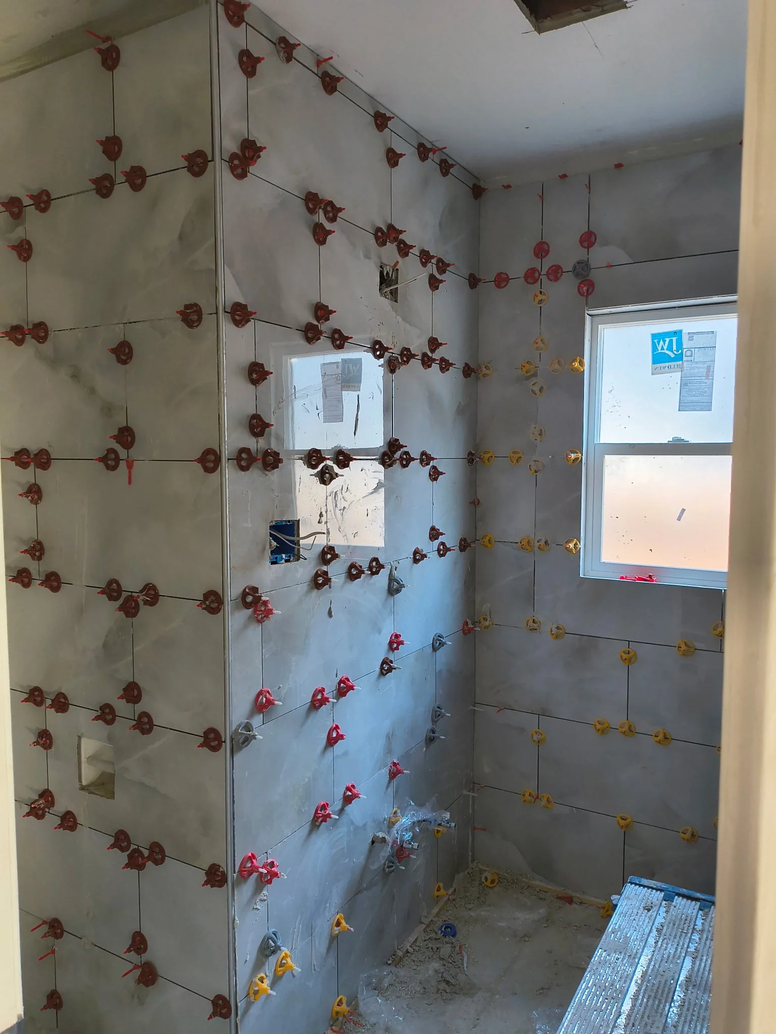 A room under construction with glass tiles being installed on the walls. Red, yellow, and silver tile spacers are visible between the tiles to ensure proper spacing. A window with a sticker on the glass is in the background, allowing natural light into the room.