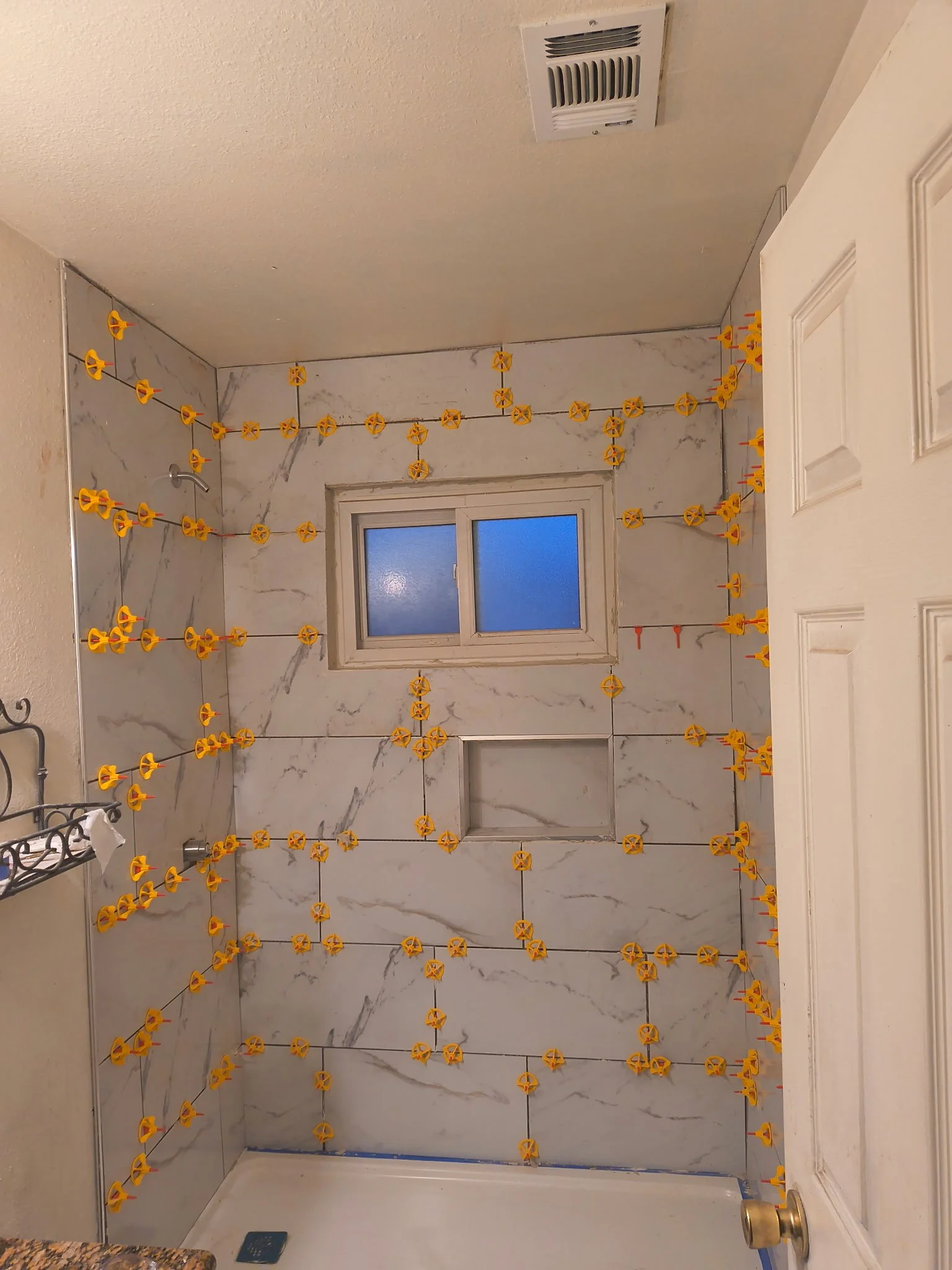 A bathroom shower area under renovation with yellow plastic clips securing a clear plastic sheet to the marble tiles. The scene also includes a small frosted window, a ceiling vent, and part of a white door on the right.