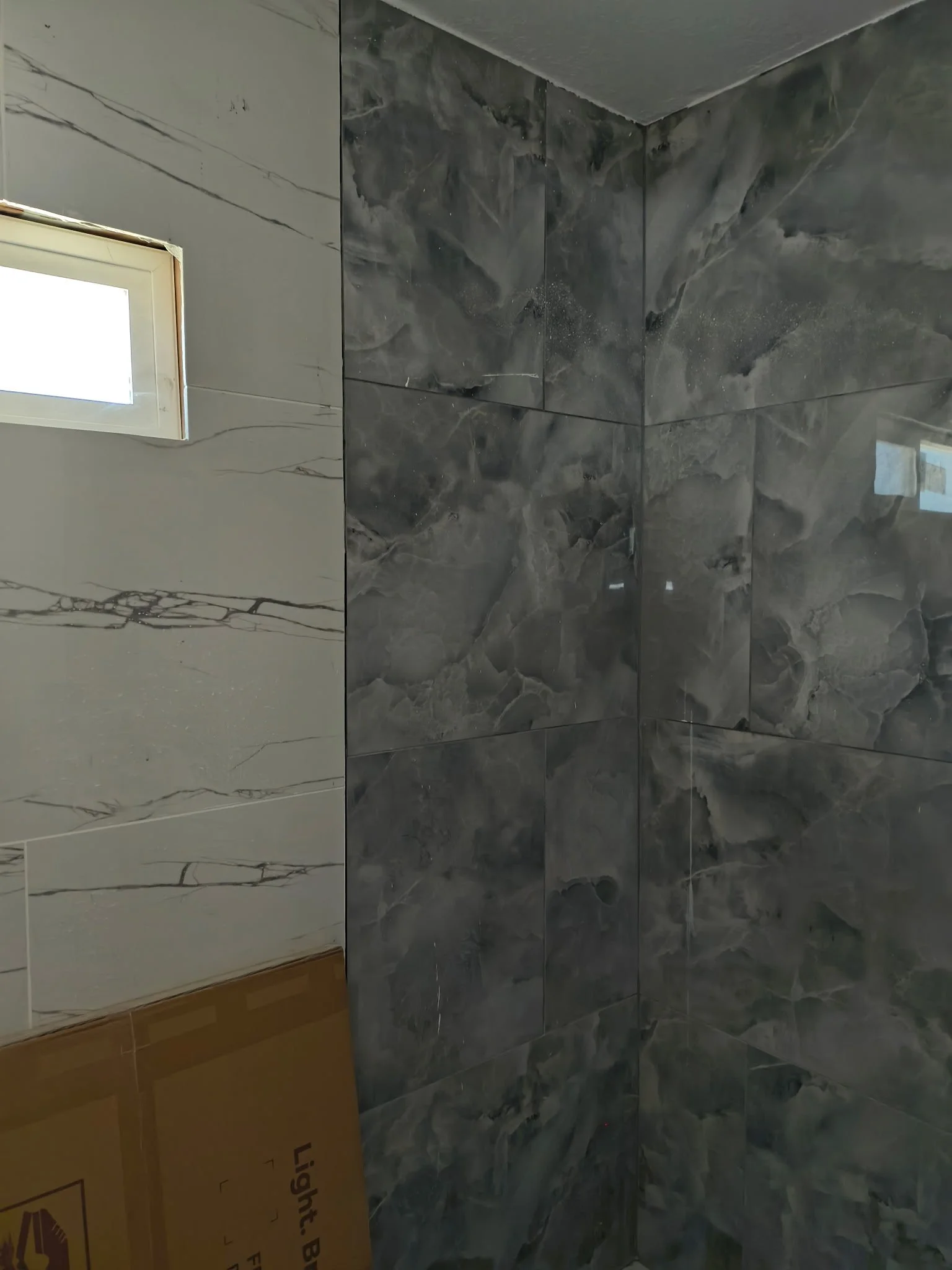 Interior corner of a room with marble tiles on the walls. The left wall has white marble with black veins, and there is a small window at the top left corner. The right wall has dark gray marble tiles with lighter gray and white veining. There is also a cardboard box on the left bottom corner.