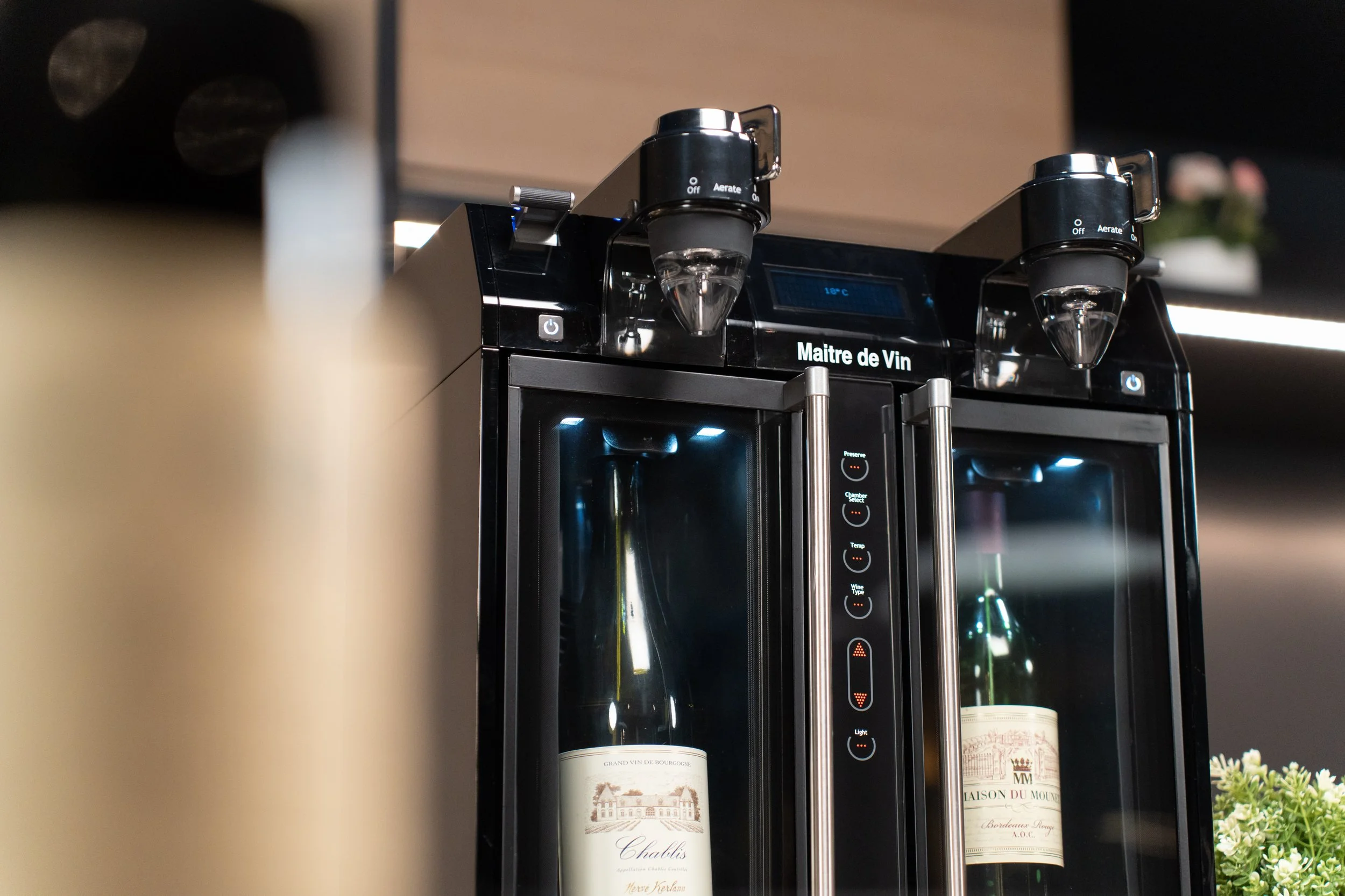 A wine dispenser with two bottles of wine, labeled 'Chablis' and 'Maison du Moulins', inside a wine fridge. The dispenser has temperature and light control buttons.
