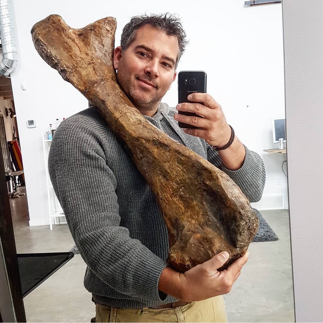Man taking a selfie in a mirror while holding a large, fossilized tree trunk or wood piece.