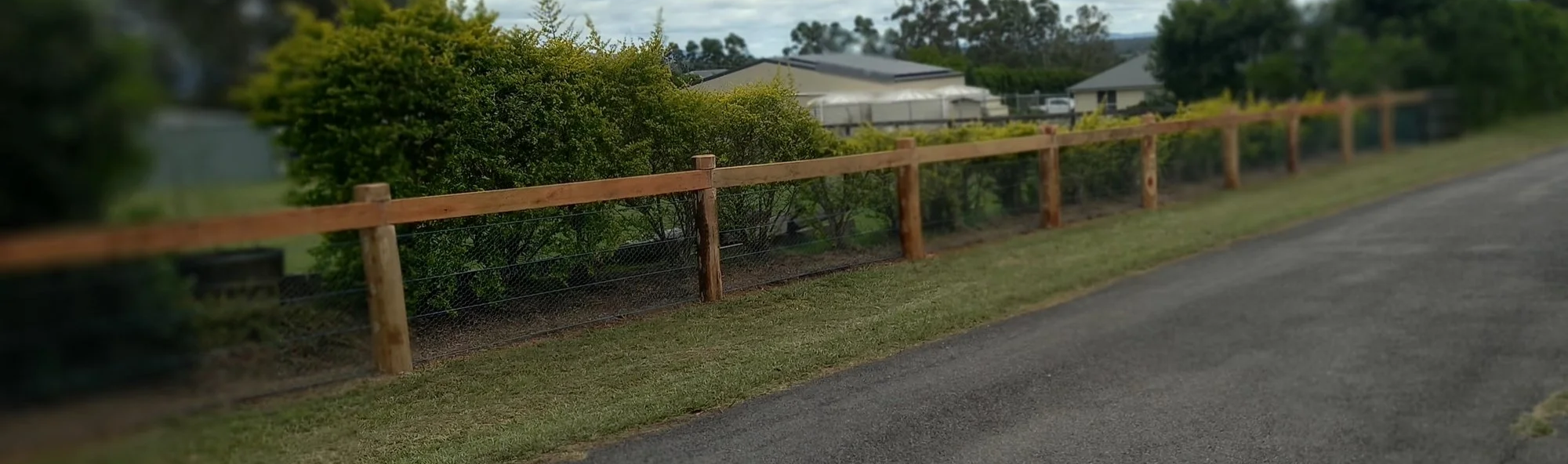 A wooden fence bordering a grassy area along a paved road, with bushes and trees behind the fence, and houses in the background under a partly cloudy sky.