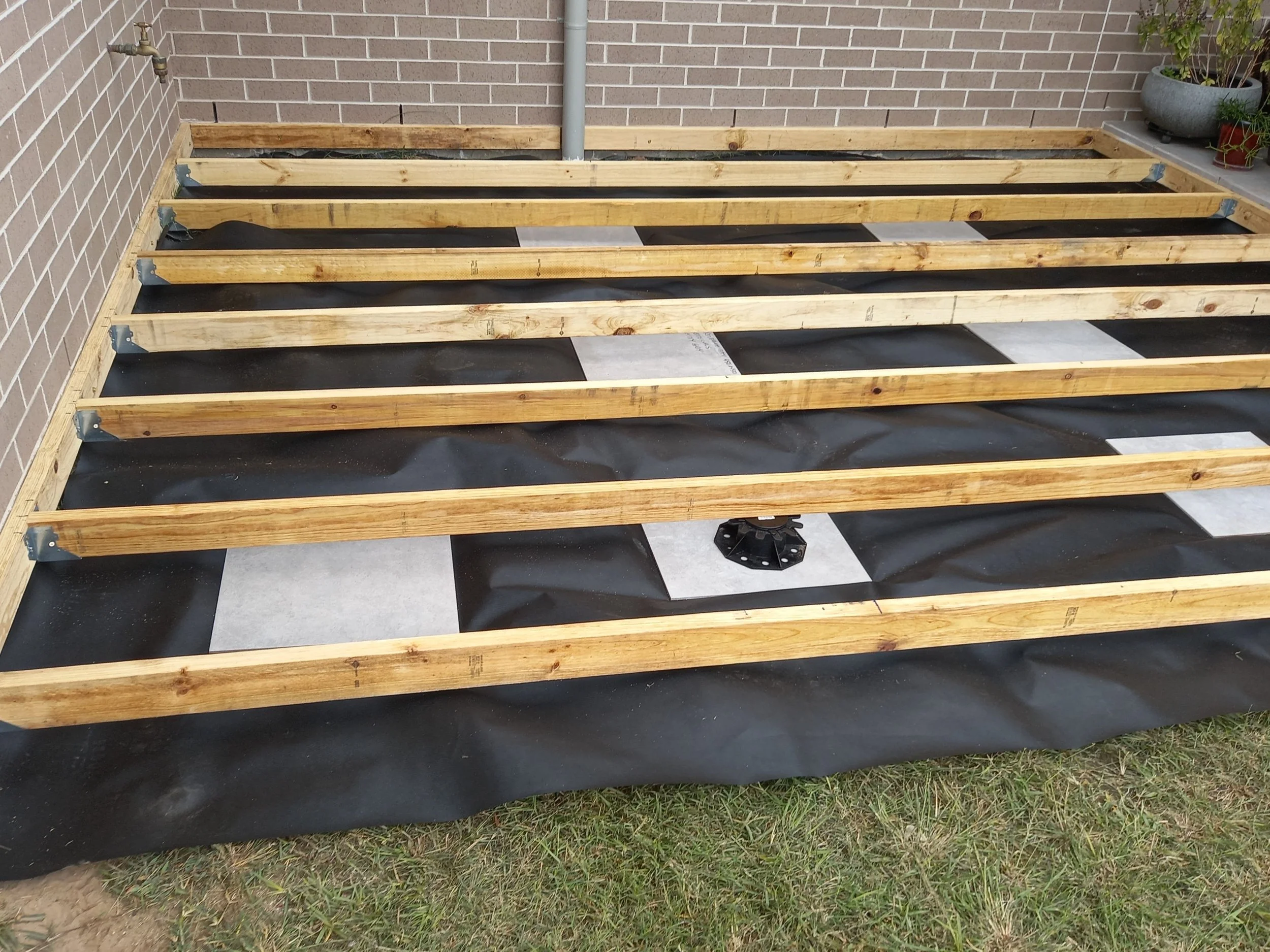 Wooden framing for a deck or platform construction against a brick wall with potted plants on the right, outdoors.