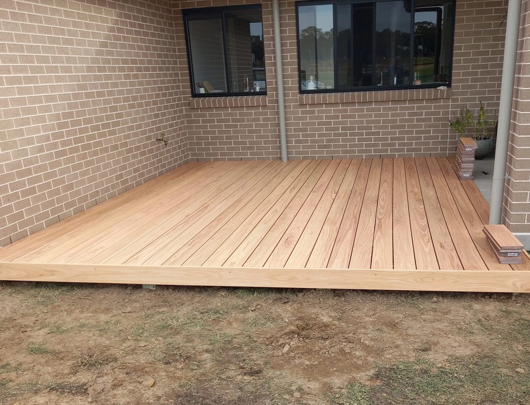 A newly built wooden deck attached to a brick house with black framed windows. The deck is off the ground, with some soil and grass visible underneath.