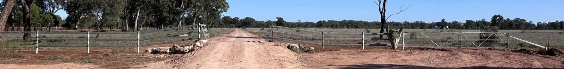 A rural dirt road with a barrier gate, flanked by wire fencing, leading towards a flat open landscape with sparse trees under a clear blue sky.