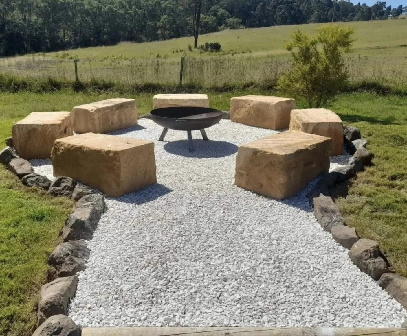 A circular fire pit with six large stone seats arranged around it on a gravel surface in an open grassy field with trees in the background.