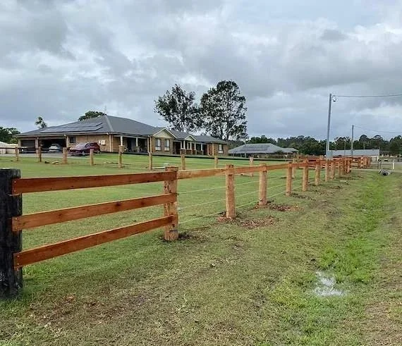 A fenced grassy yard in front of a house with a porch and several cars parked outside. The fence is made of wooden posts and rails, stretching into the distance under cloudy skies.