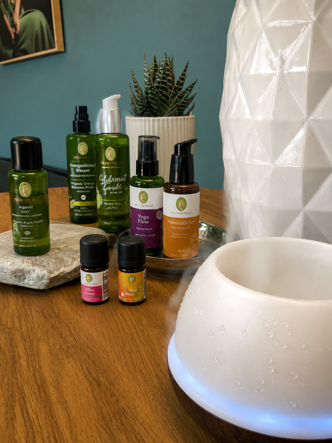 Aromatherapie-Set mit ätherischen Ölen, einem Räuchergefäß mit Rauch, einer Pflanze in einer weißen Topf, einem Holztablett, einem Stein und einer Wand mit einem Gemälde im Hintergrund.