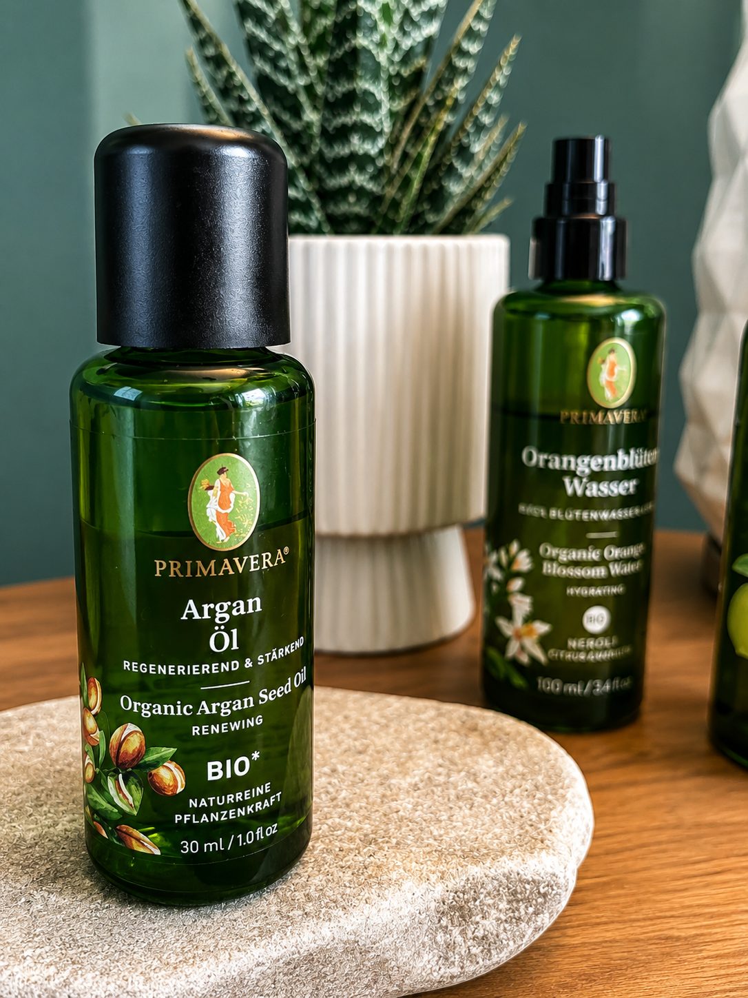 Grüne Flasche mit schwarzem Deckel und Etikett mit dem Text "Primavera Argan Öl" und einem Bild von Argankernen, neben einer weiteren Flasche "Orangenblüten Wasser" auf einem Holztisch. Im Hintergrund eine weiße Pflanzentopf mit einer grünen Pflanze.