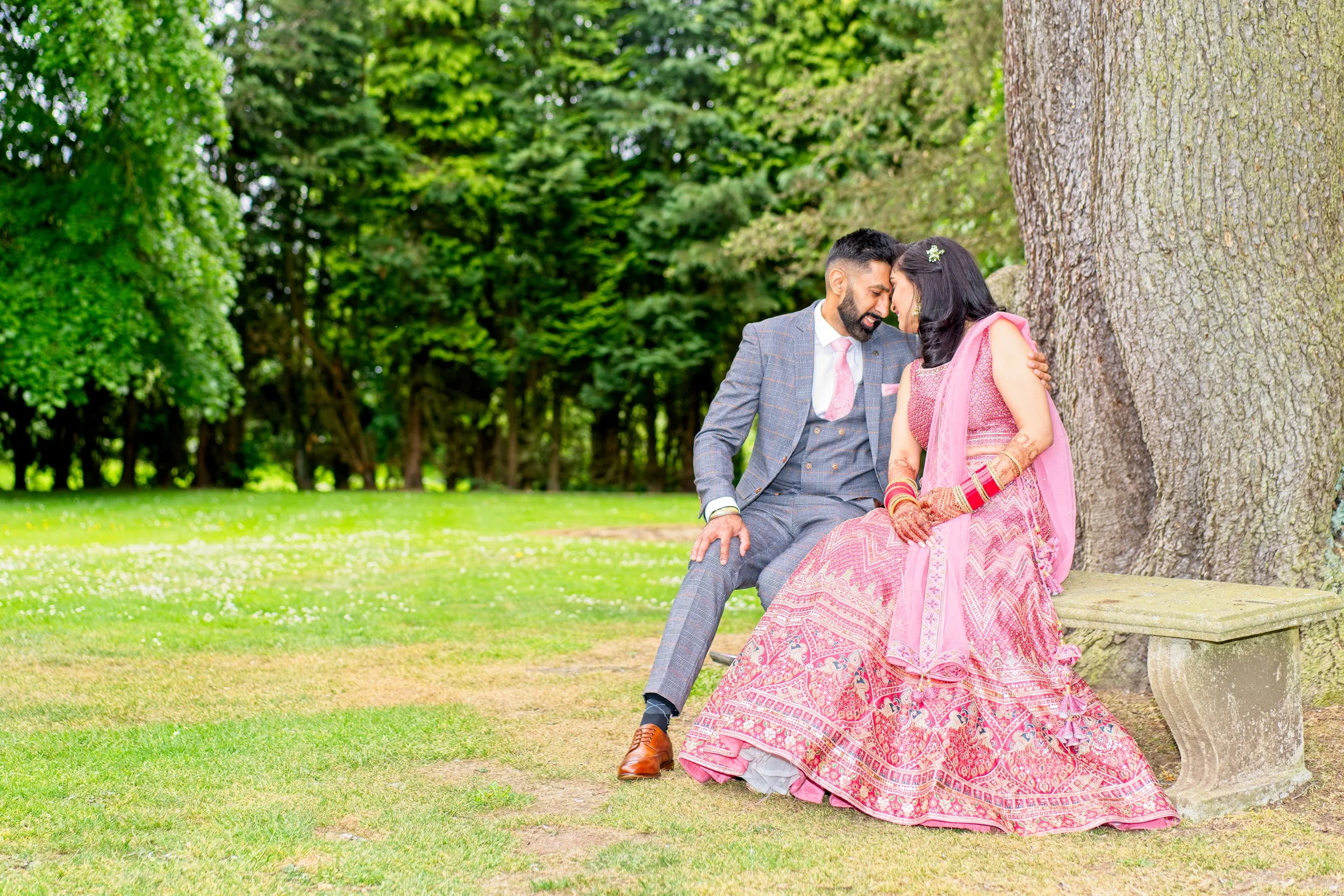 Man and woman dressed in wedding attire sitting on a park bench under a large tree, leaning heads together, smiling and sharing a romantic moment at MacDonald's Hotel, Coventry. Photography by photographer based in Southam, Warwickshire