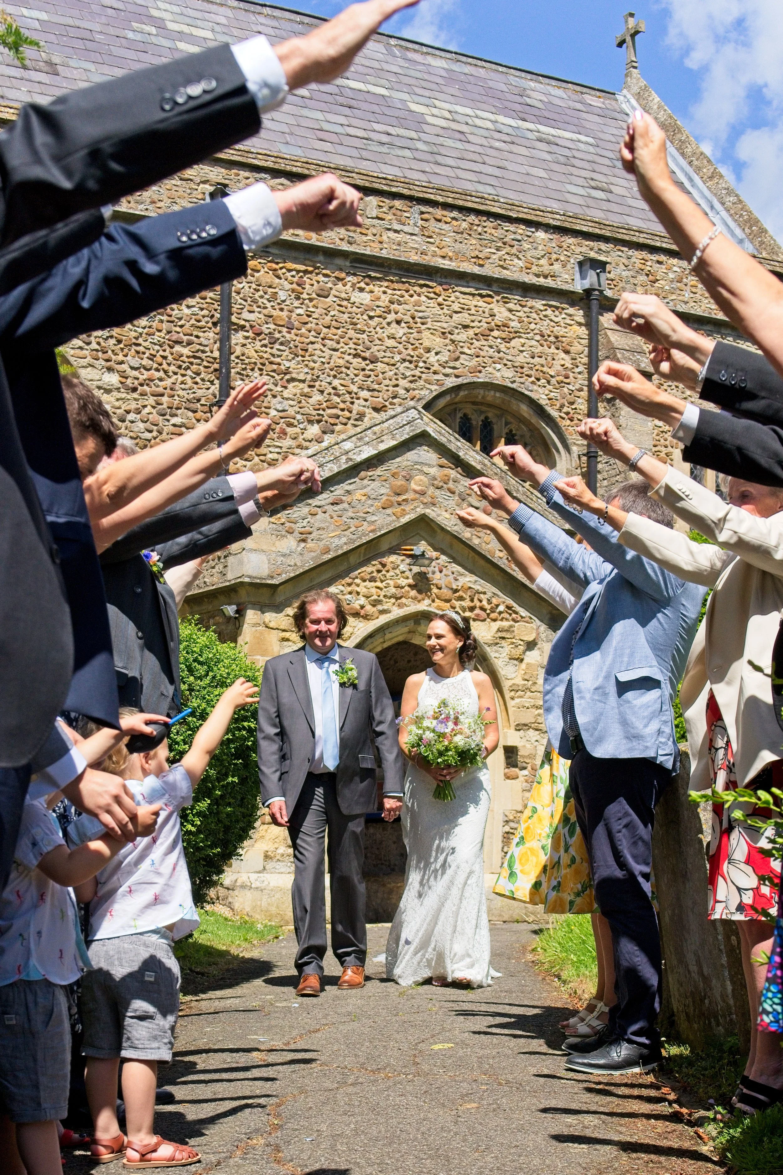 Bride holding a bouquet walking down the aisle with her father at a wedding ceremony outside a stone church, surrounded by guests raising their arms in celebration. Photography by photographer based in Southam, Warwickshire