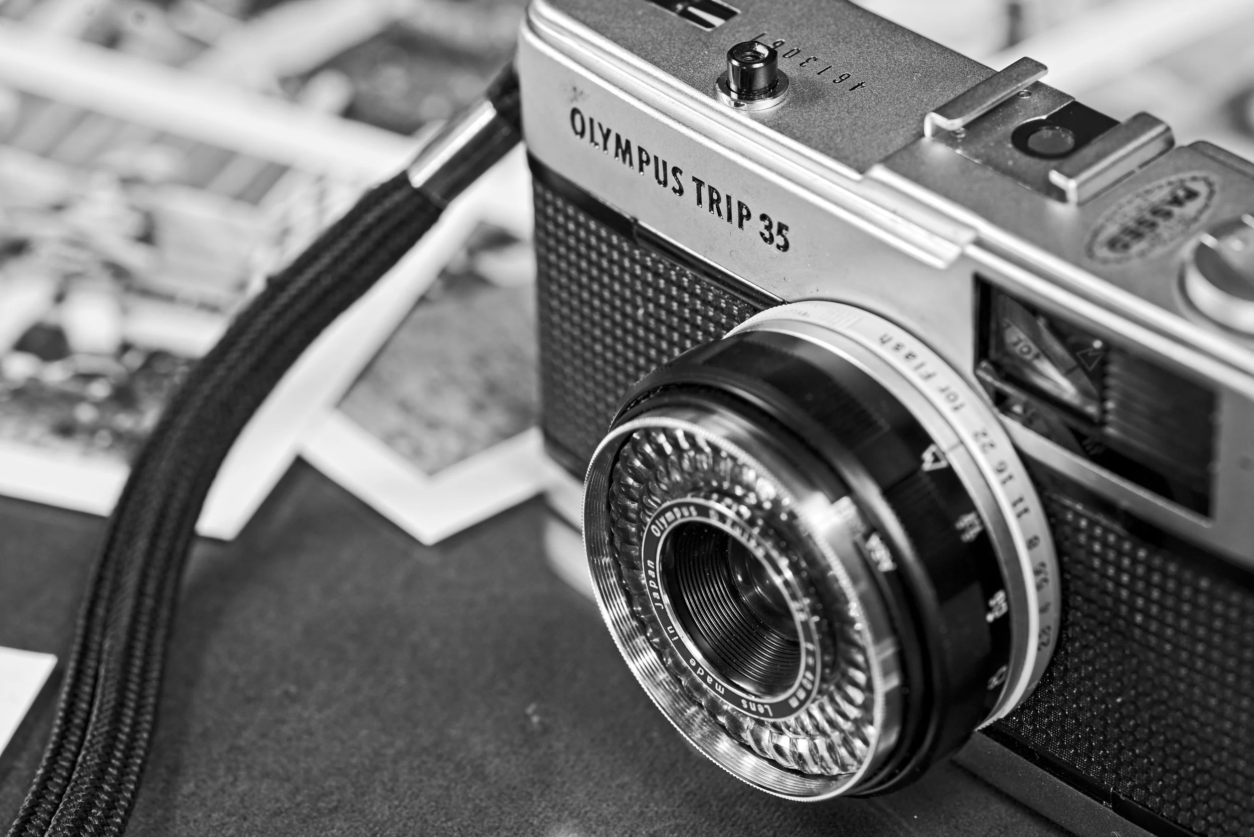 A black and white photo of an Olympus Trip 35 camera with a lens, strap, and some printed photos in the background. Photography by photographer based in Southam, Warwickshire