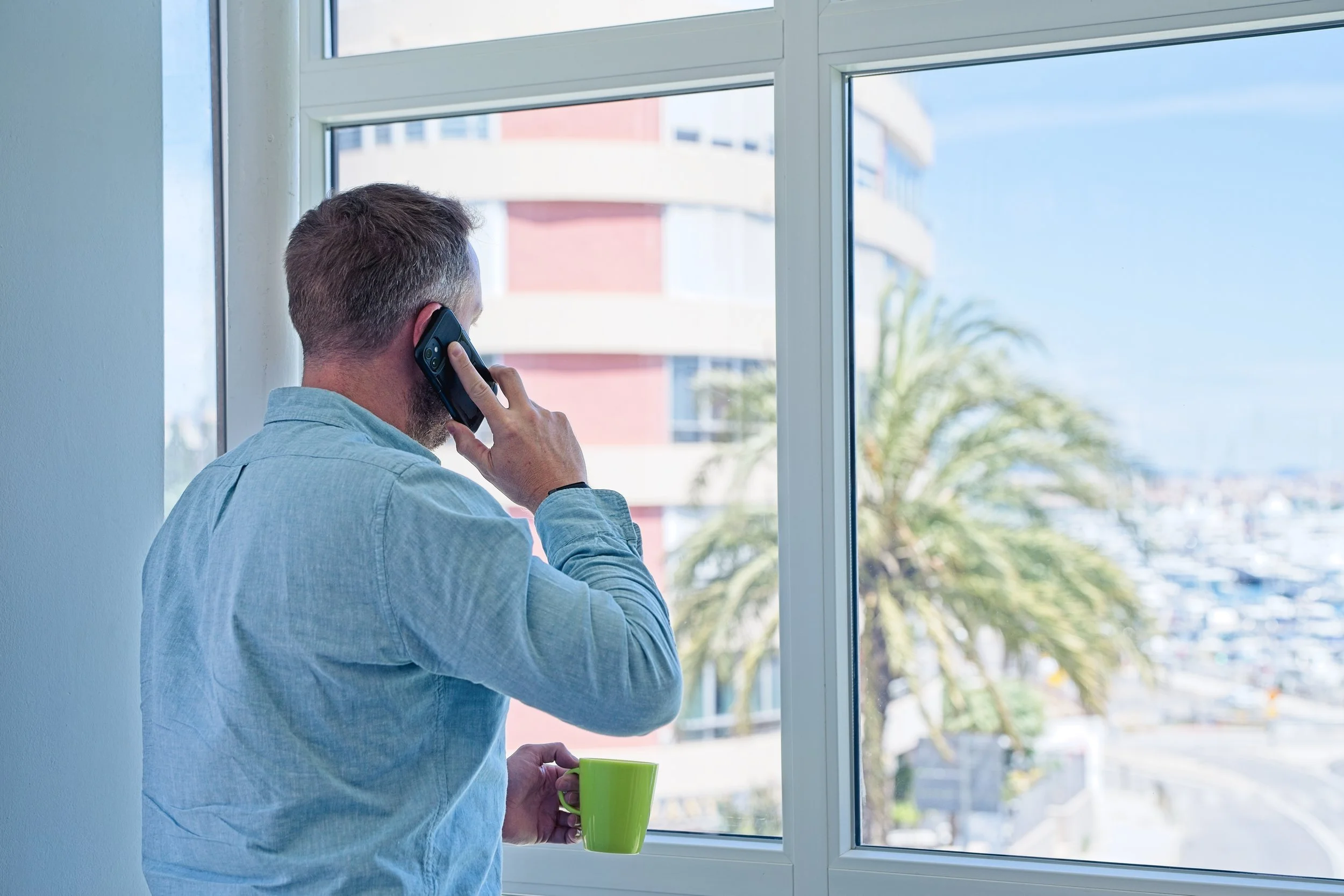 Man standing by window, talking on cell phone, holding a green mug. Branding photography Warwickshire.