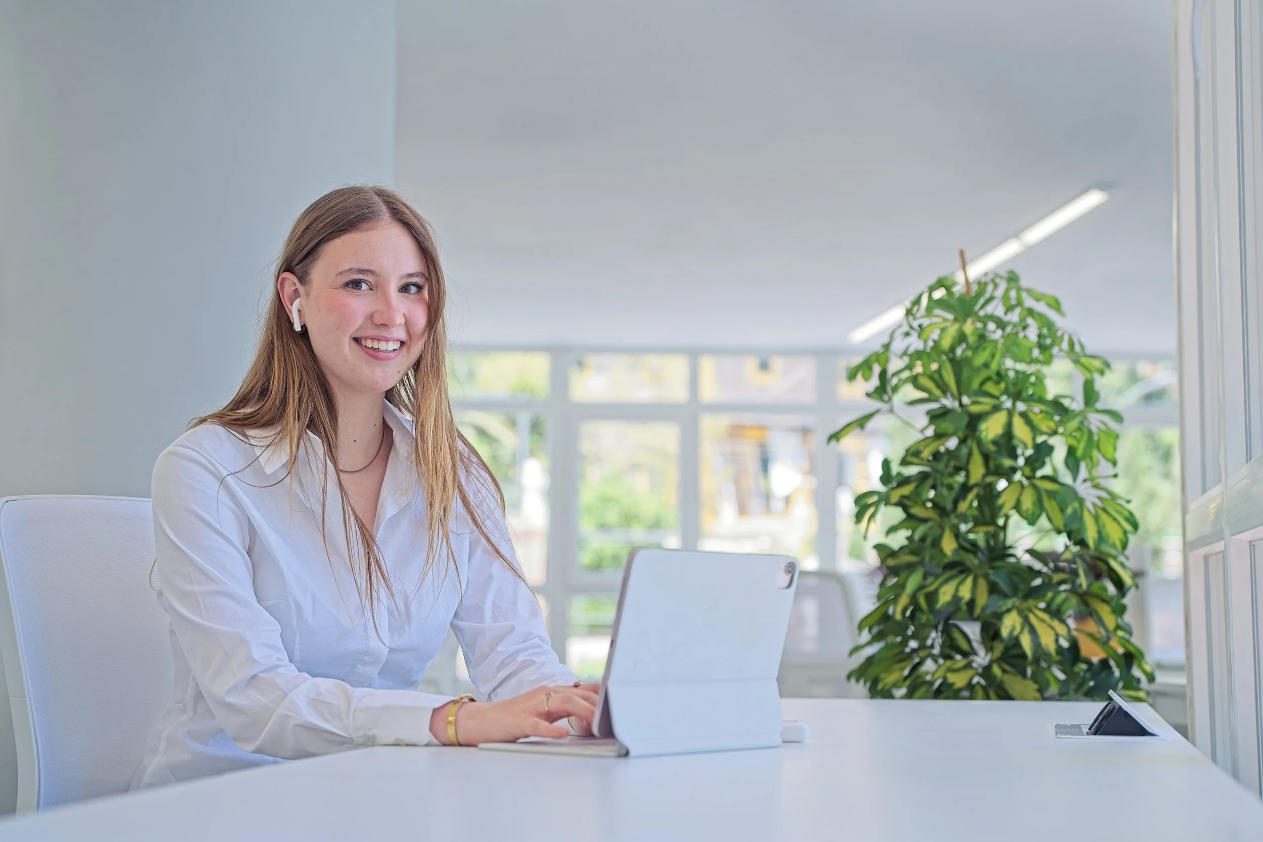 A young woman with long red hair, wearing a white shirt and AirPods, sitting at a white desk with a tablet in a bright, modern office. Branding and corporate photography in Warwickshire.