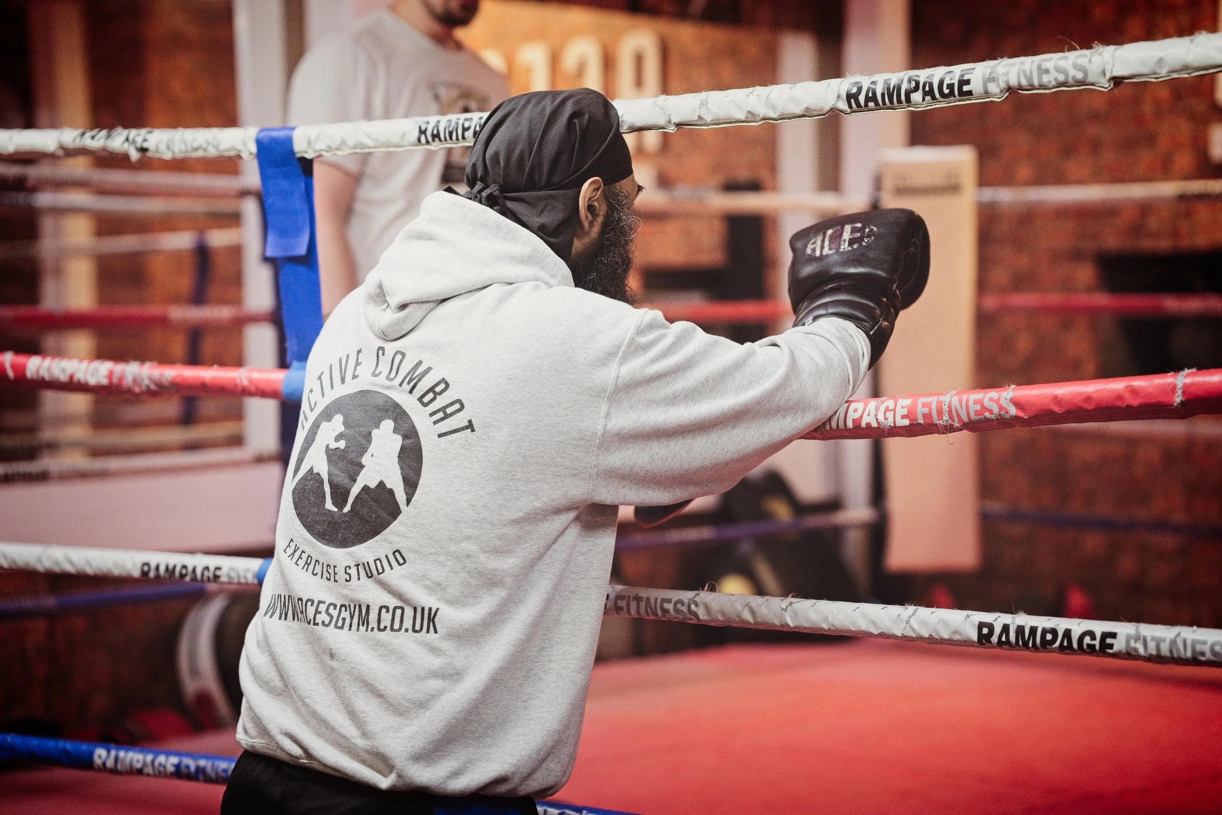 A man practicing boxing in a gym ring, wearing boxing gloves and a gray hoodie with 'Active Combat Exercise Studio' logo on the back, with a trainer standing nearby. Warwickshire sports photography.