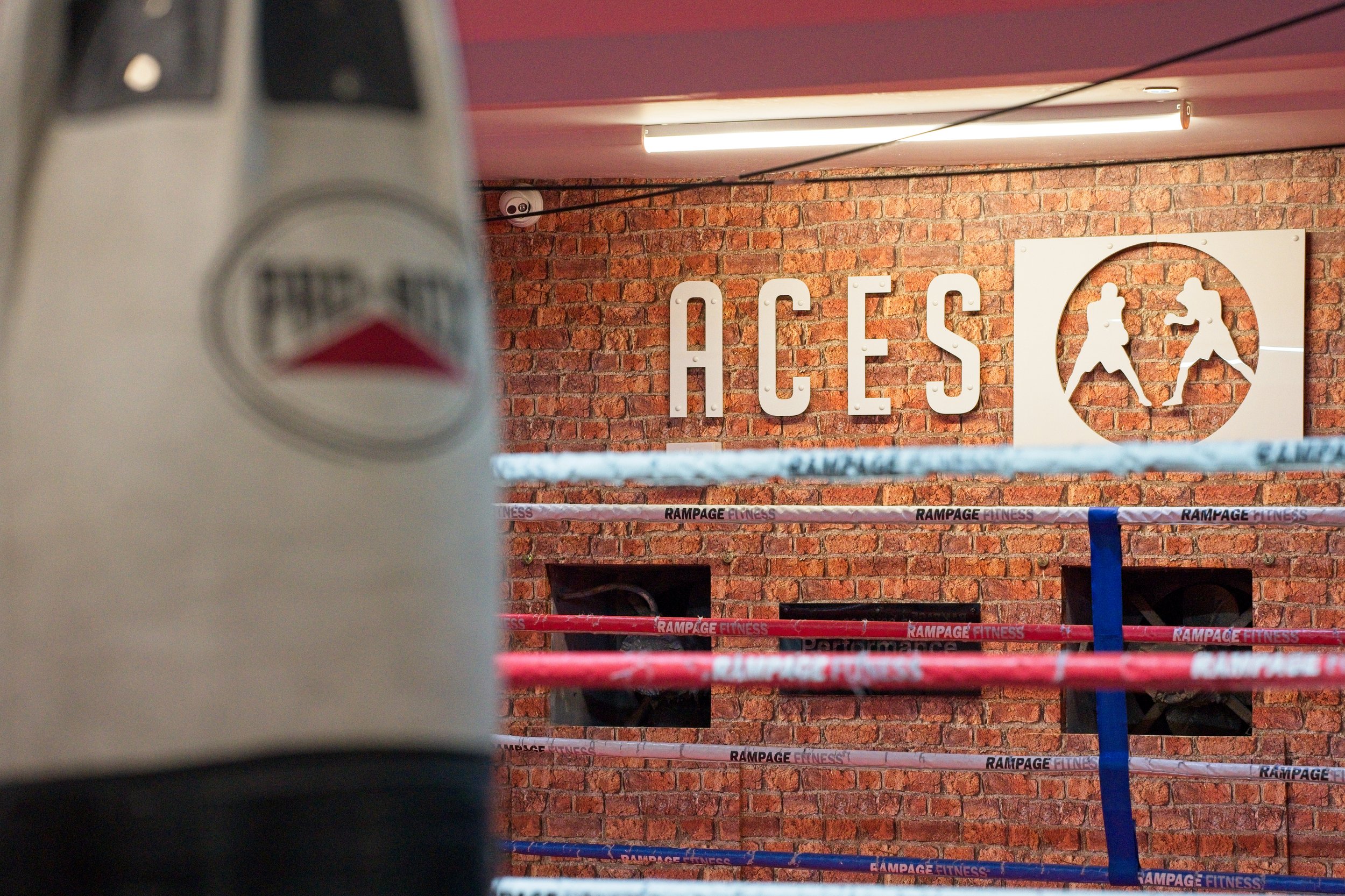 A boxing gym with a brick wall, a large 'ACES' sign, and boxing ring ropes in the foreground. Warwickshire sports photography.
