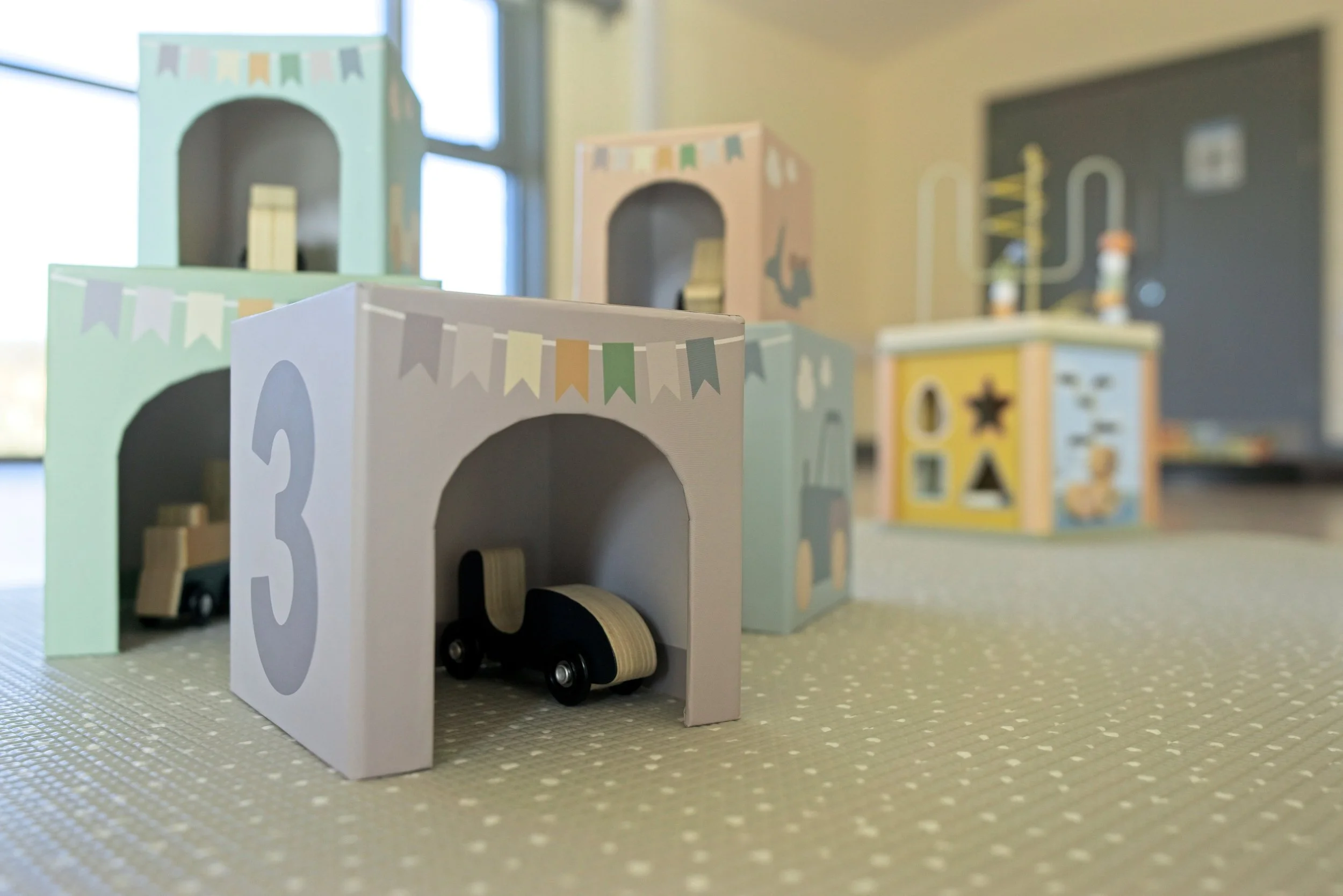 Colorful wooden toy garages numbered 2 and 3 with small toy cars inside, set on a textured surface with a window in the background. Warwickshire branding photography.