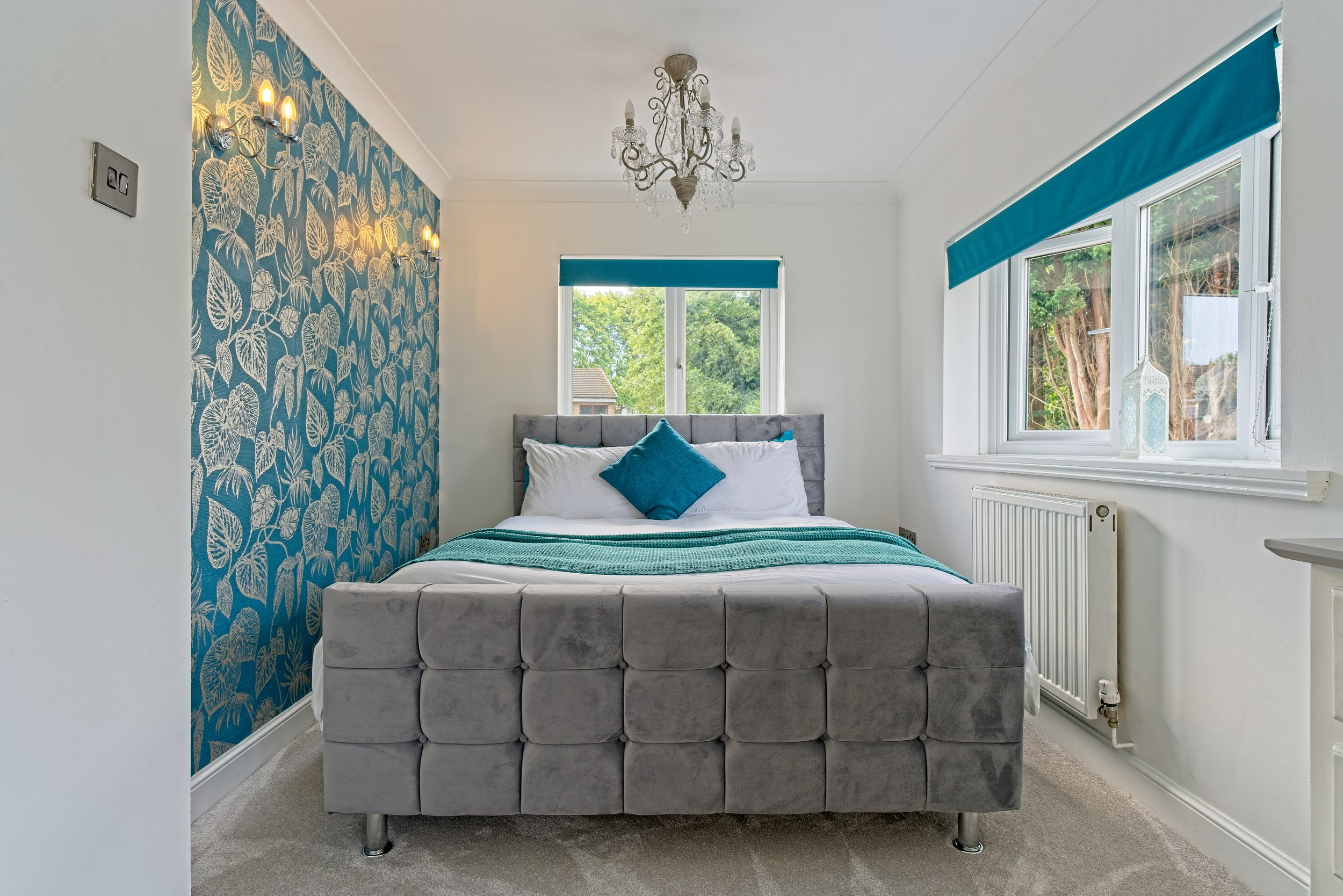 A bedroom with a bed, decorative wall with blue and gold leaf pattern, chandelier, and large windows with blue blinds showing quality photography by Southam based Photographer