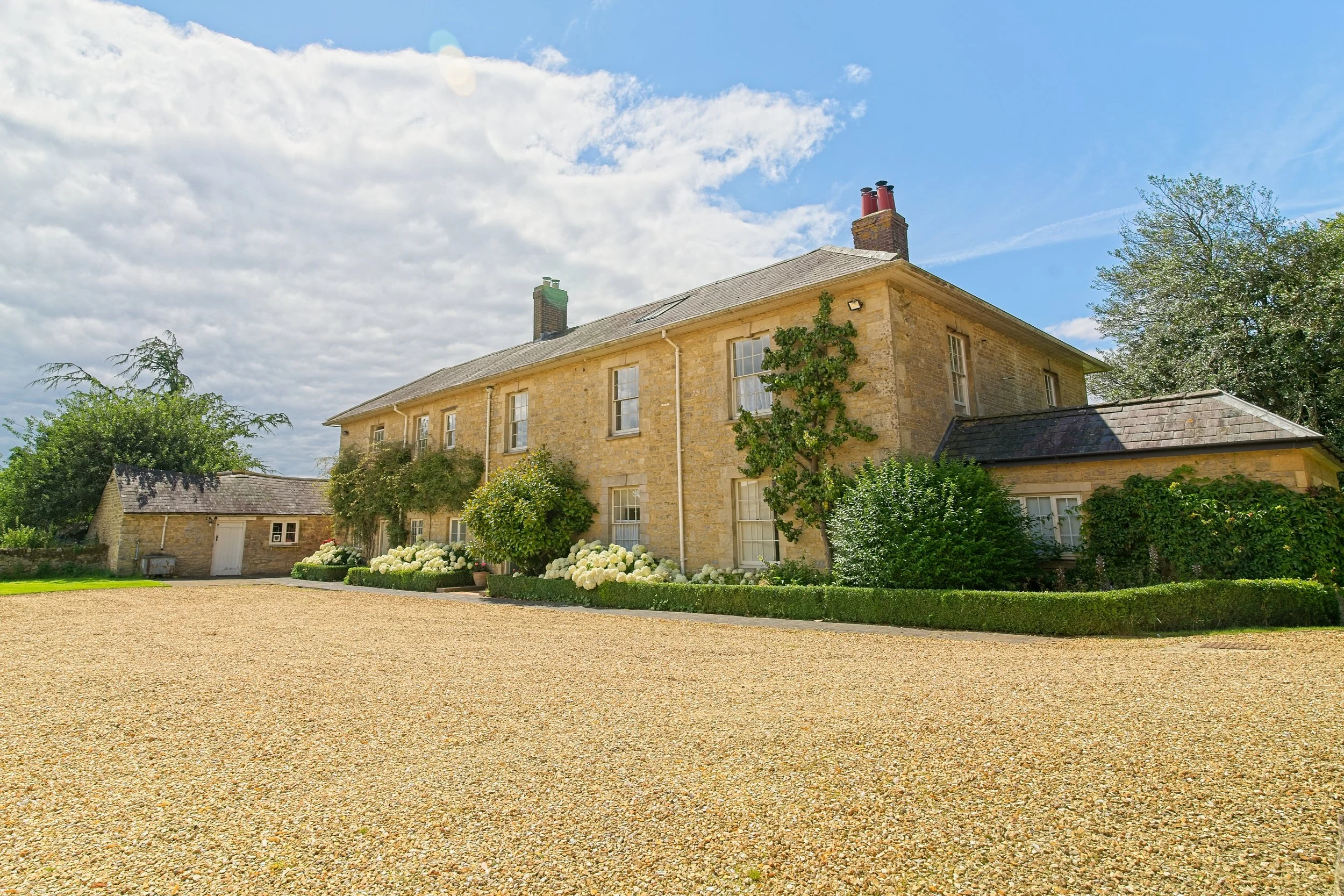 A large, two-story stone house with multiple windows and a sloped roof, surrounded by greenery and a gravel driveway. Real estate photography in the Cotswolds and Warwickshire.