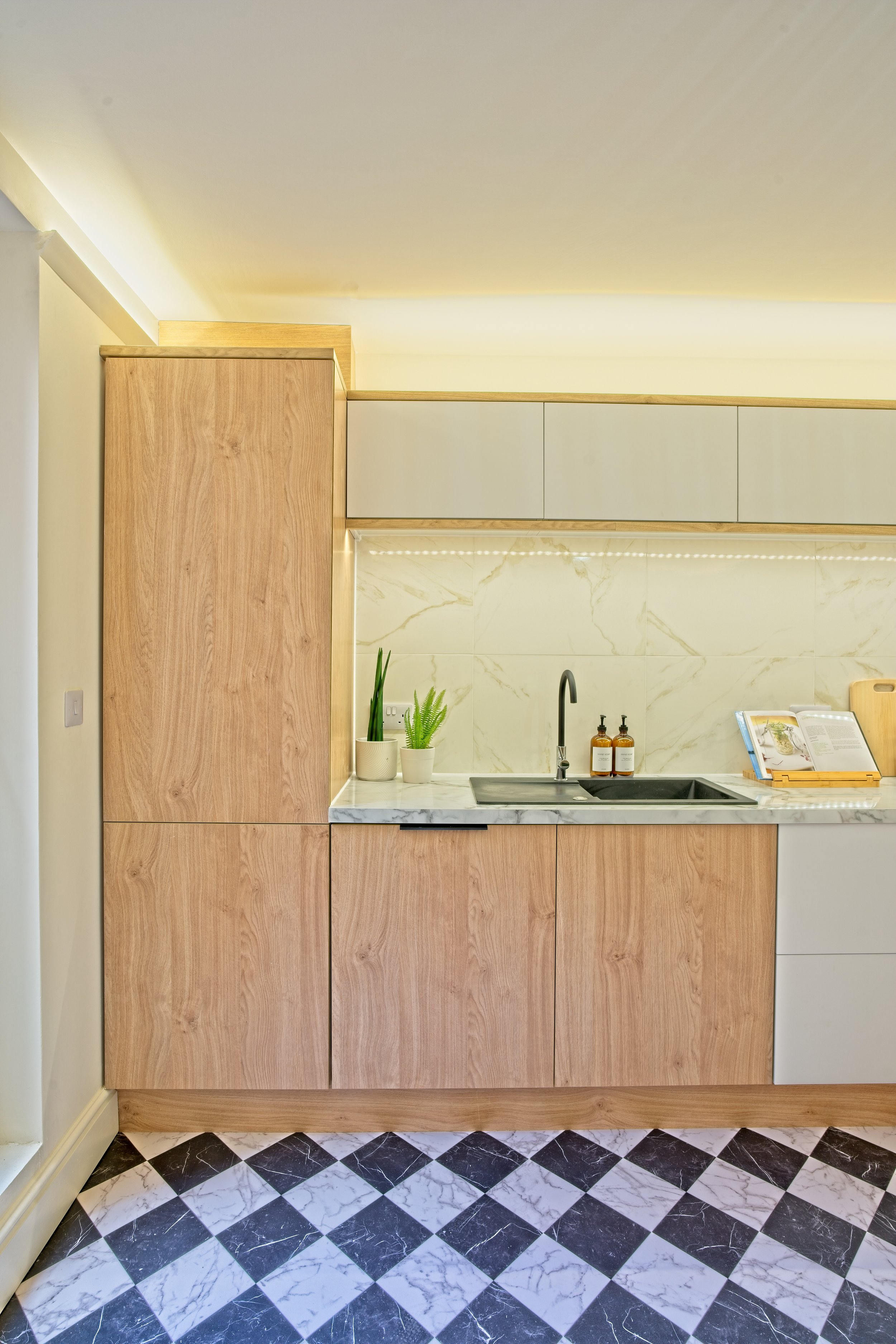 Modern kitchen with light wood cabinets, marble backsplash, black sink, two hand soap bottles, potted plants, open cookbook on a stand, and a patterned black and white tile floor. Photography by photographer based in Southam, Warwickshire