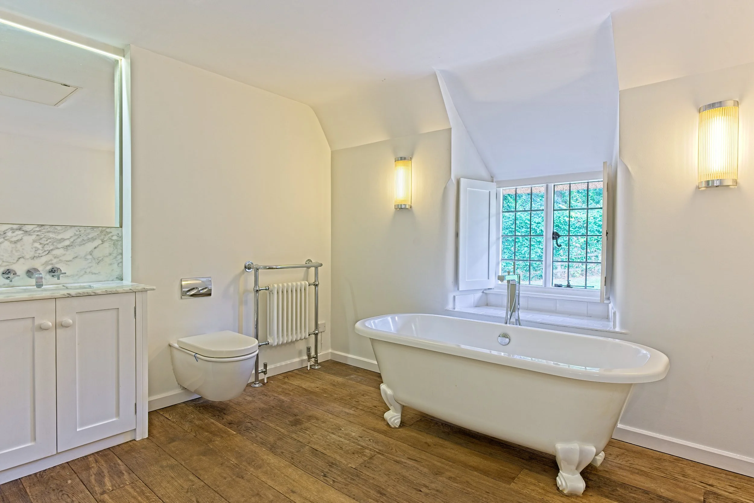 Bathroom with a clawfoot tub, a toilet, a small sink, wooden flooring, and a window with shutters overlooking greenery. Property Photography by photographer based in Southam, Warwickshire