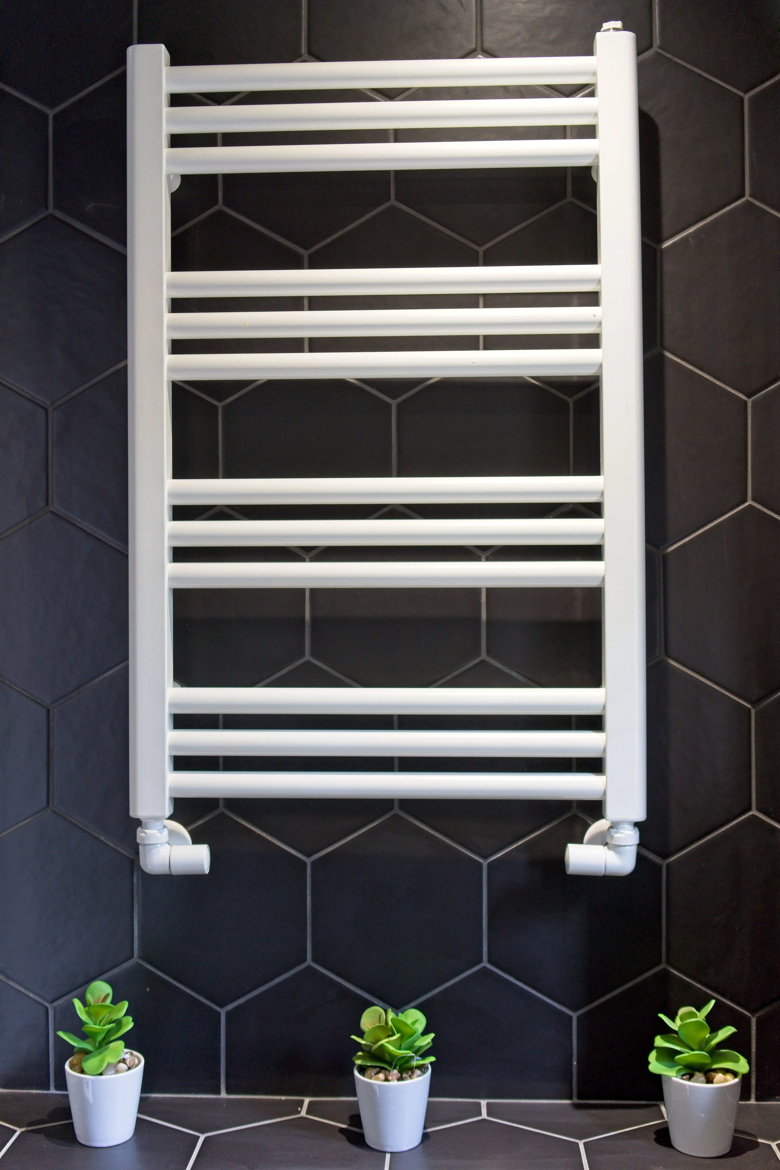 White towel radiator mounted on a black tiled wall with three small green potted plants at the base. Photography by photographer based in Southam, Warwickshire