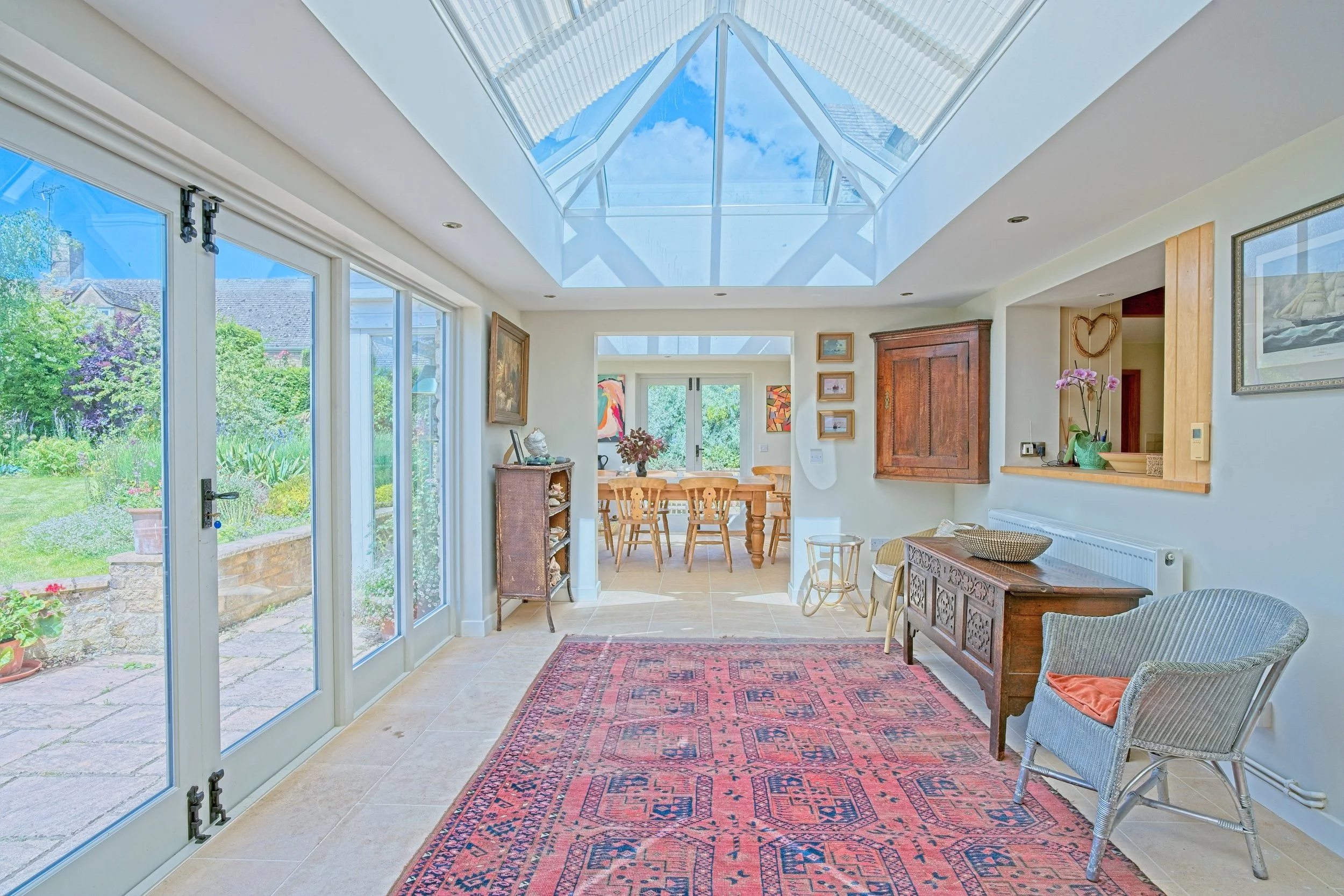Bright living room with large glass sliding doors, a skylight ceiling, and a view of a garden outside. Contains a red patterned rug, wicker chairs, wooden furniture, and artwork on the walls. Photography by photographer based in Southam, Warwickshire