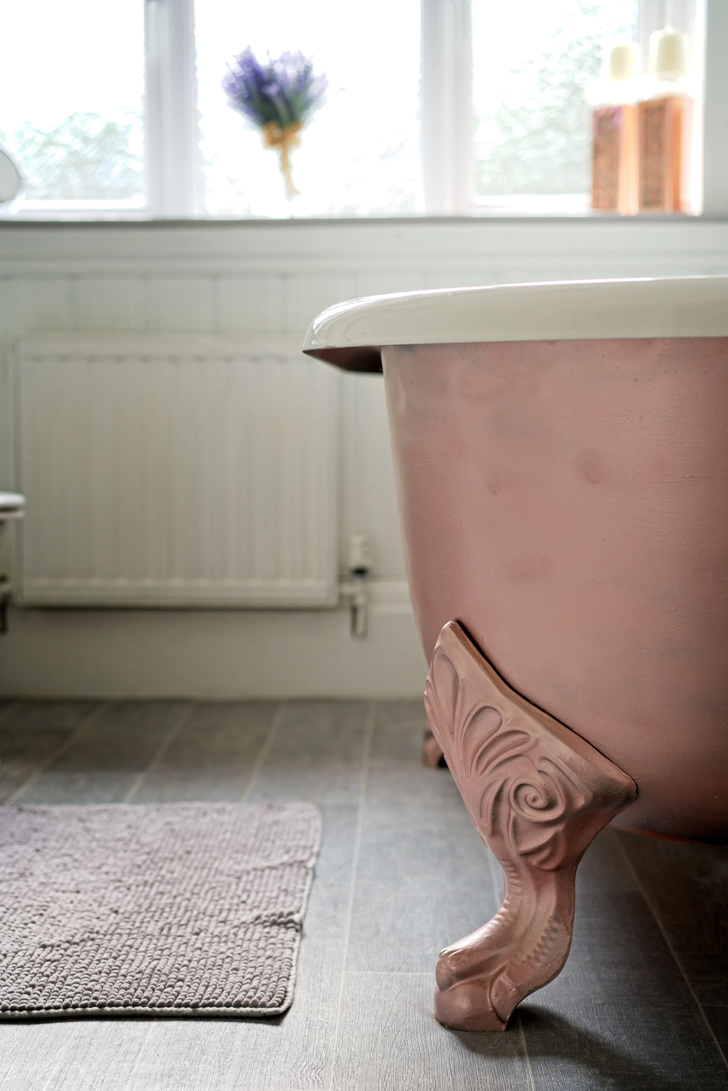 Close-up of a vintage pink clawfoot bathtub in a bathroom with a window showing a purple flower bouquet and white candles in the background. Property Photography by photographer based in Southam, Warwickshire