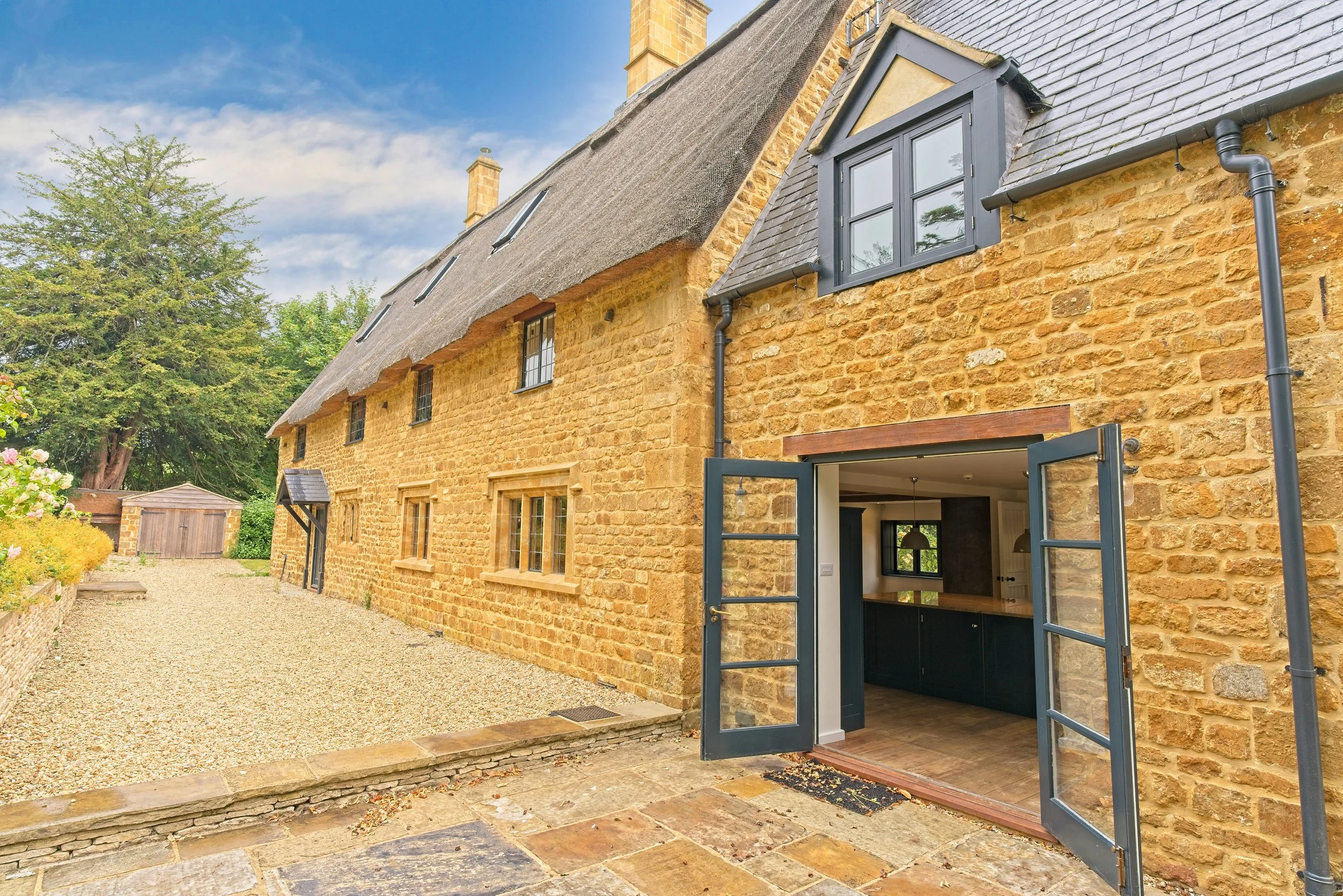 View of a traditional stone-built house with an open door leading into a kitchen, surrounded by a gravel pathway and lush greenery, under a blue sky. Photography by photographer based in Southam, Warwickshire