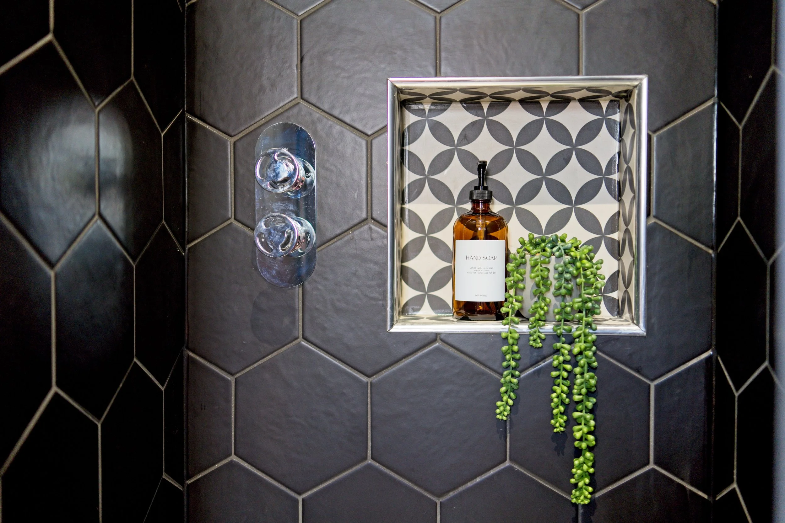 Black tiled shower wall with built-in shelf holding a hand soap bottle, a small green succulent plant, and a patterned background. Property photography by photographer based in Southam, Warwickshire
