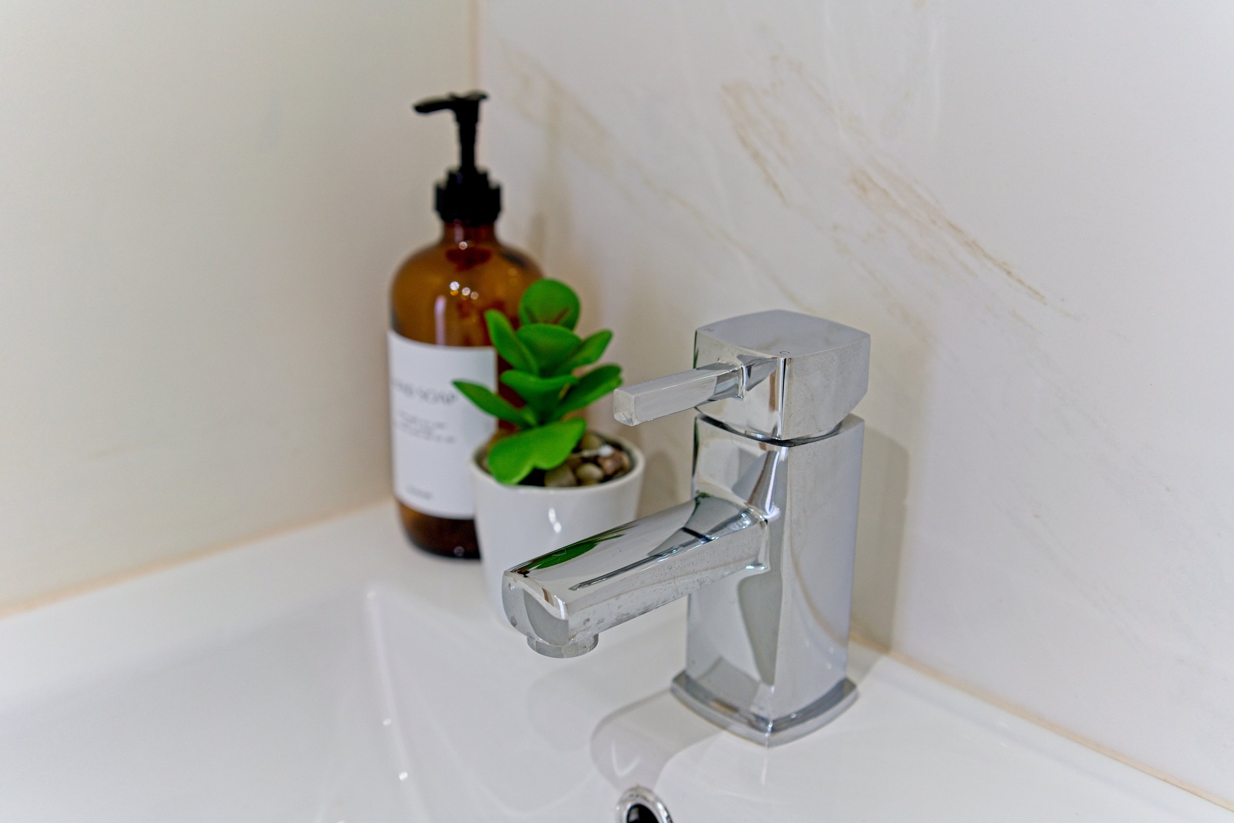 Bathroom sink with a modern chrome faucet, a potted succulent plant, and a brown bottle with a pump dispenser on a beige tiled wall. Photography by photographer based in Southam, Warwickshire