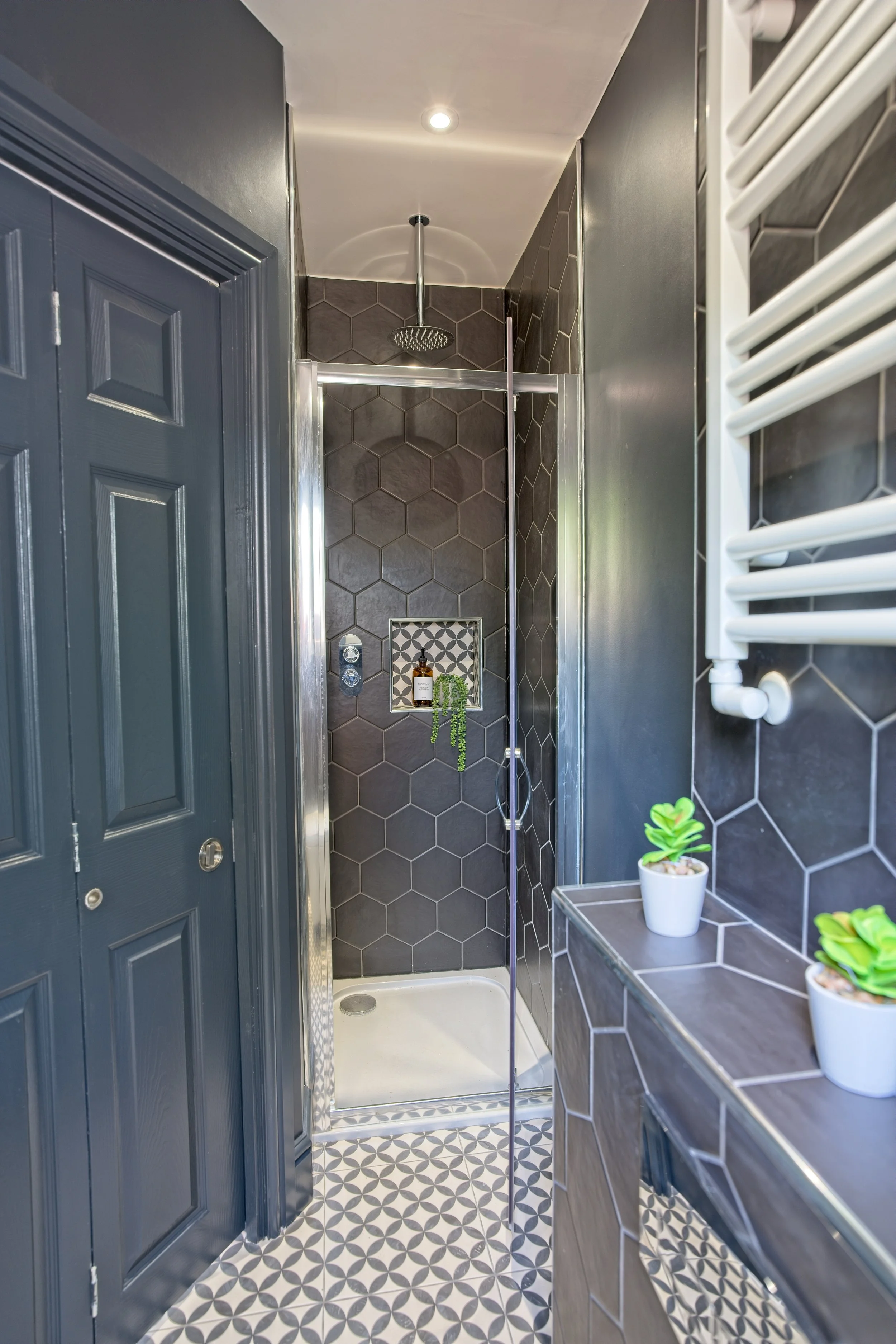 A modern shower with dark grey hexagon tiles, a glass door, a built-in niche for toiletries, black and white geometric patterned floor tiles, and small potted succulents on a ledge. Property photography by photographer based in Southam, Warwickshire