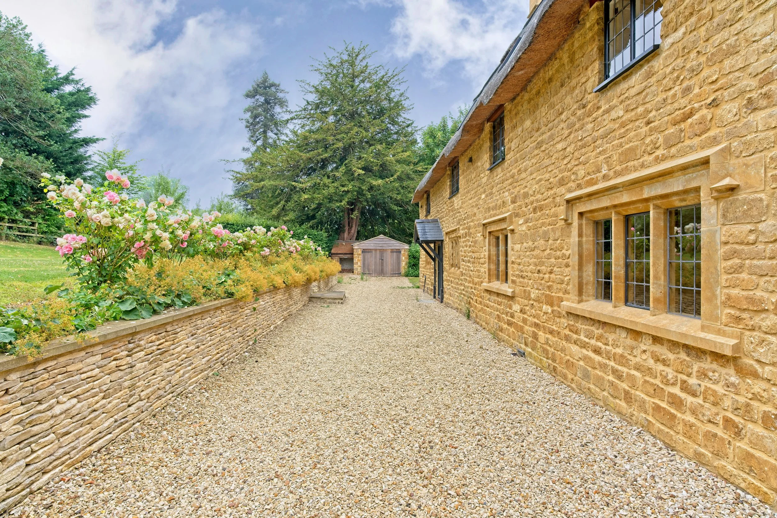 Side view of a brick house with small windows, a gravel pathway, and a garden with pink and white flowers and green trees. Property Photography by photographer based in Southam, Warwickshire