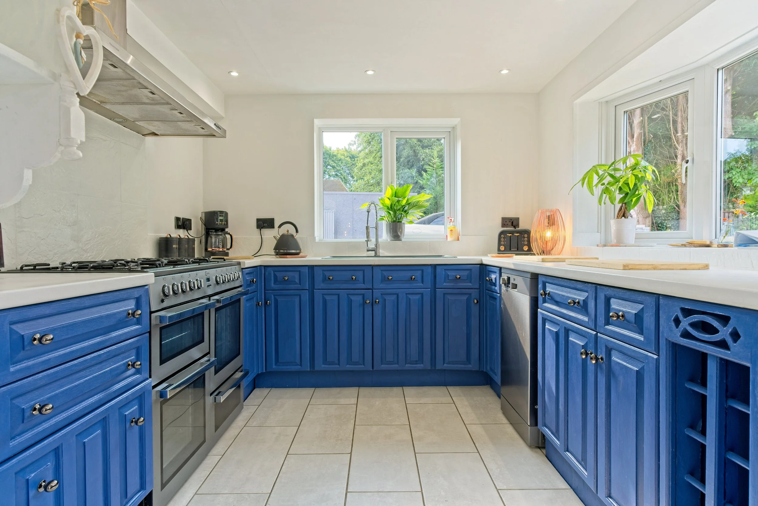 Kitchen with blue cabinets, white countertops, a window with trees outside, and various appliances including a stove, toaster, and coffee maker. Photography by photographer based in Southam, Warwickshire