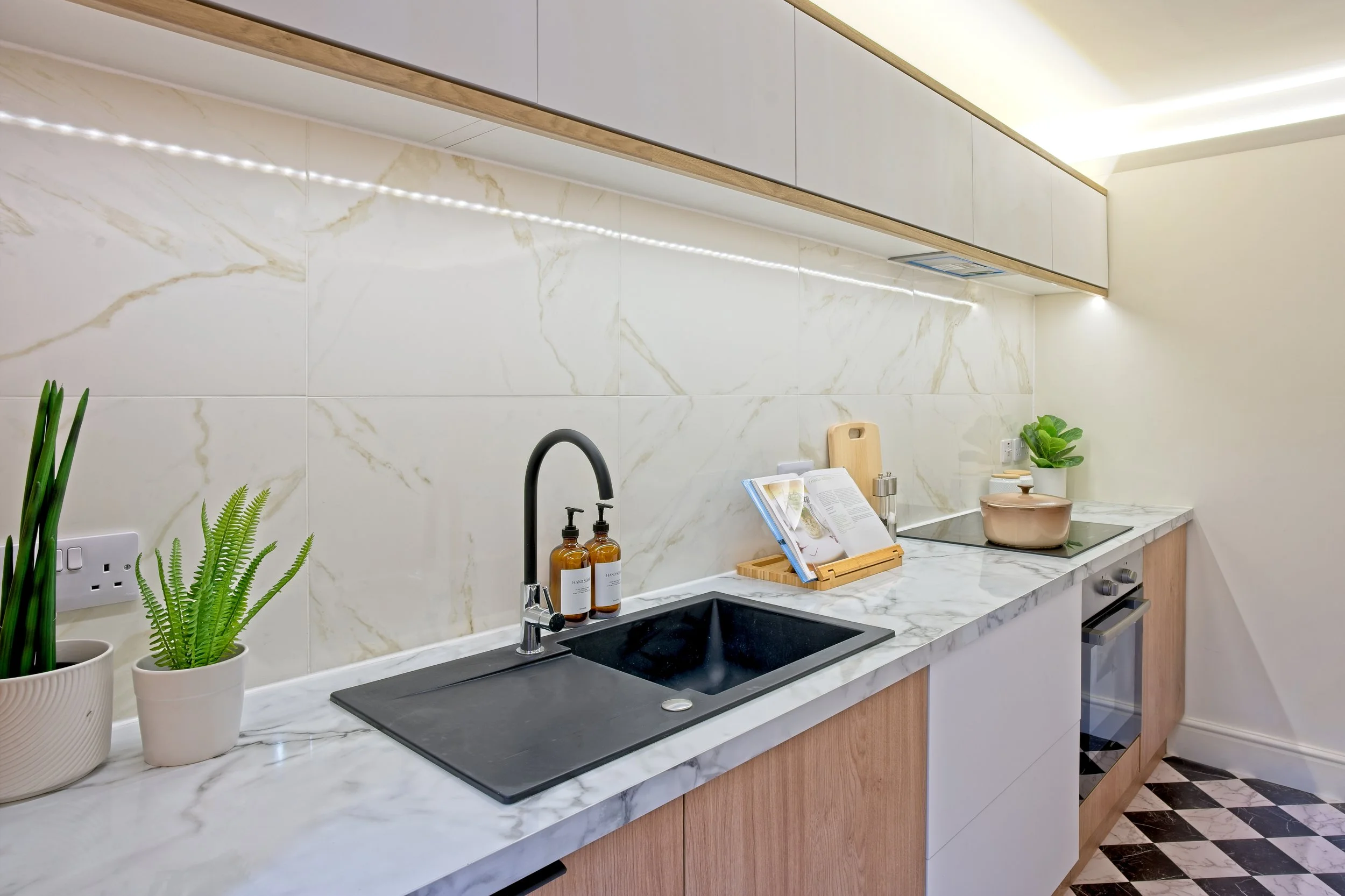 Modern kitchen with white marble countertop, black sink, wooden cabinets, potted plants, open cookbook, and stovetop. Photography by photographer based in Southam, Warwickshire