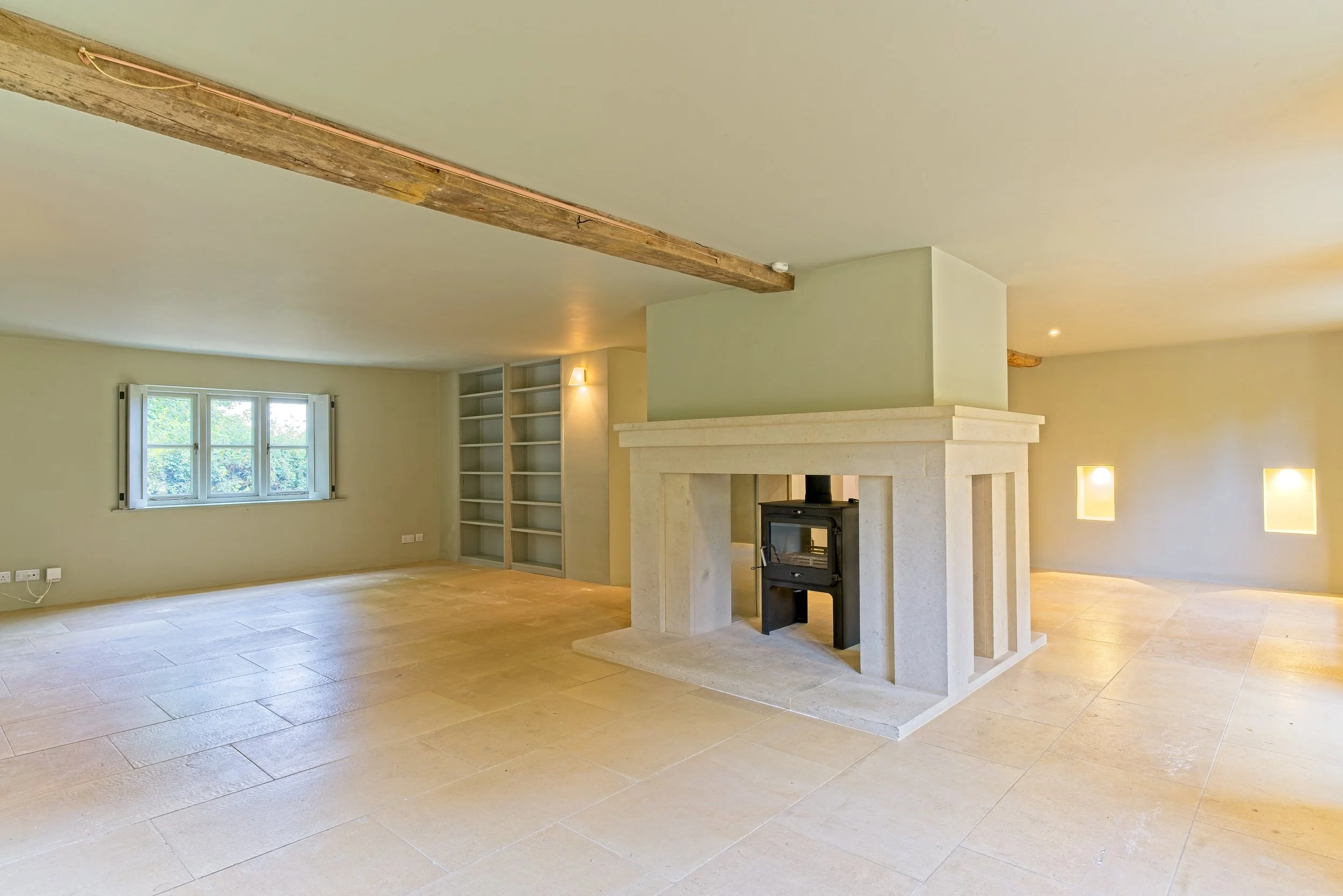 Empty living room with a stone fireplace, large window, wooden ceiling beam, built-in bookshelf, and wall niches with lighting. Property Photography by photographer based in Southam, Warwickshire