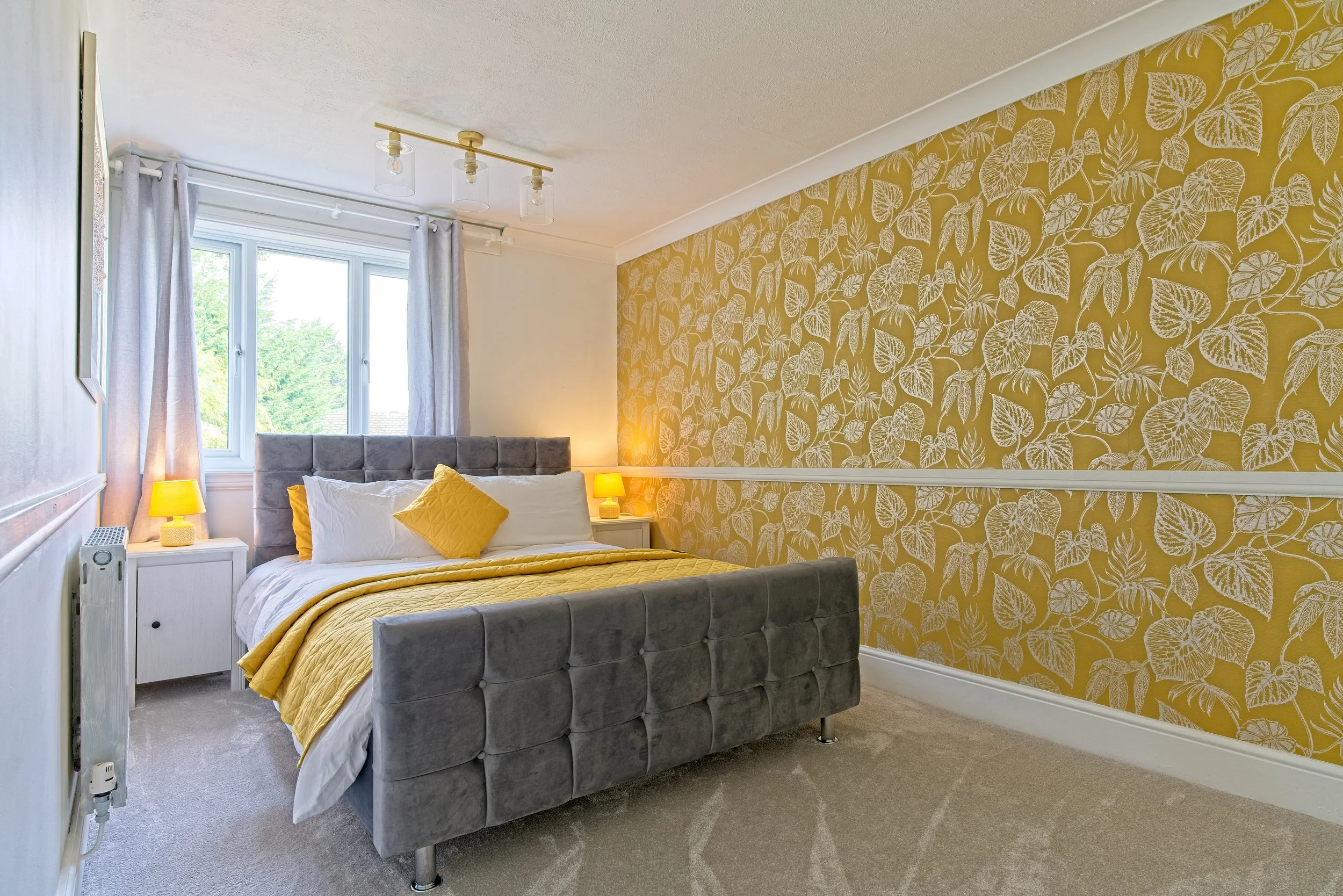 A bedroom with a gray velvet bed, yellow bedding and pillows, white nightstands with yellow lamps, a window with white curtains, and yellow leaf-patterned wallpaper on one wall. Property Photography by photographer based in Southam, Warwickshire