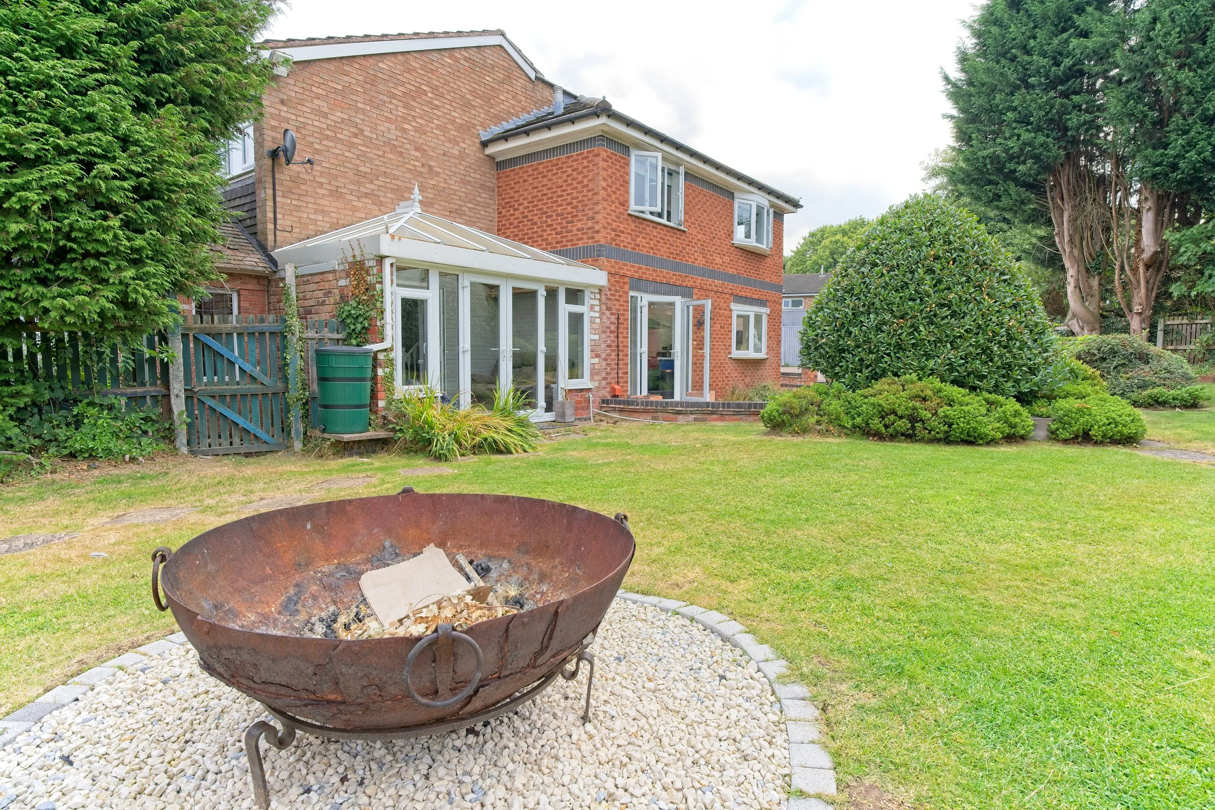 Backyard with a brick house, a grassy lawn, a rusted fire pit on gravel, and various bushes and trees. Photography by photographer based in Southam, Warwickshire
