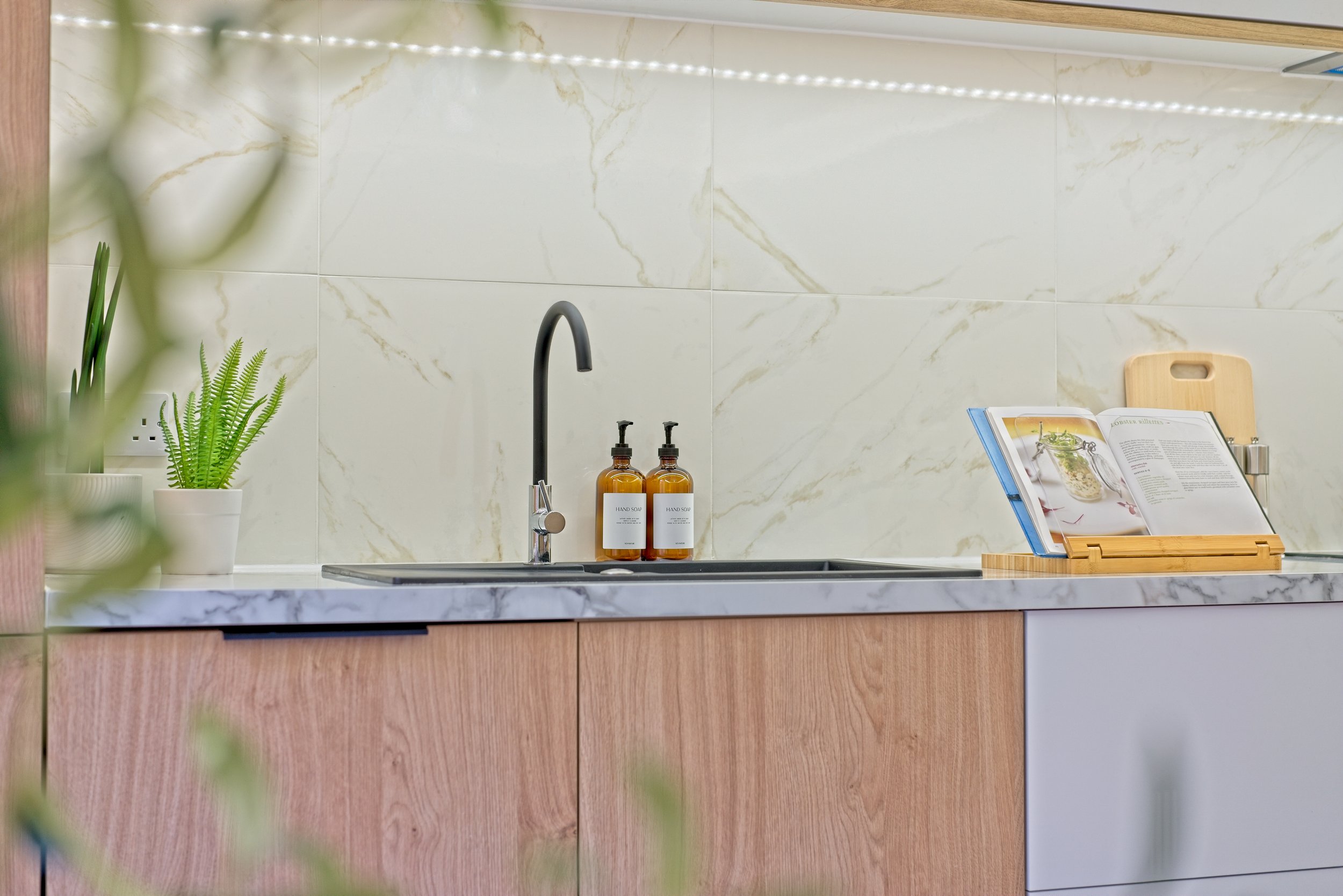 Kitchen sink with a black faucet, two amber hand soap bottles, and a wooden cookbook stand holding open cookbooks, with a potted fern and partially visible green foliage in the foreground. Photography by photographer based in Southam, Warwickshire