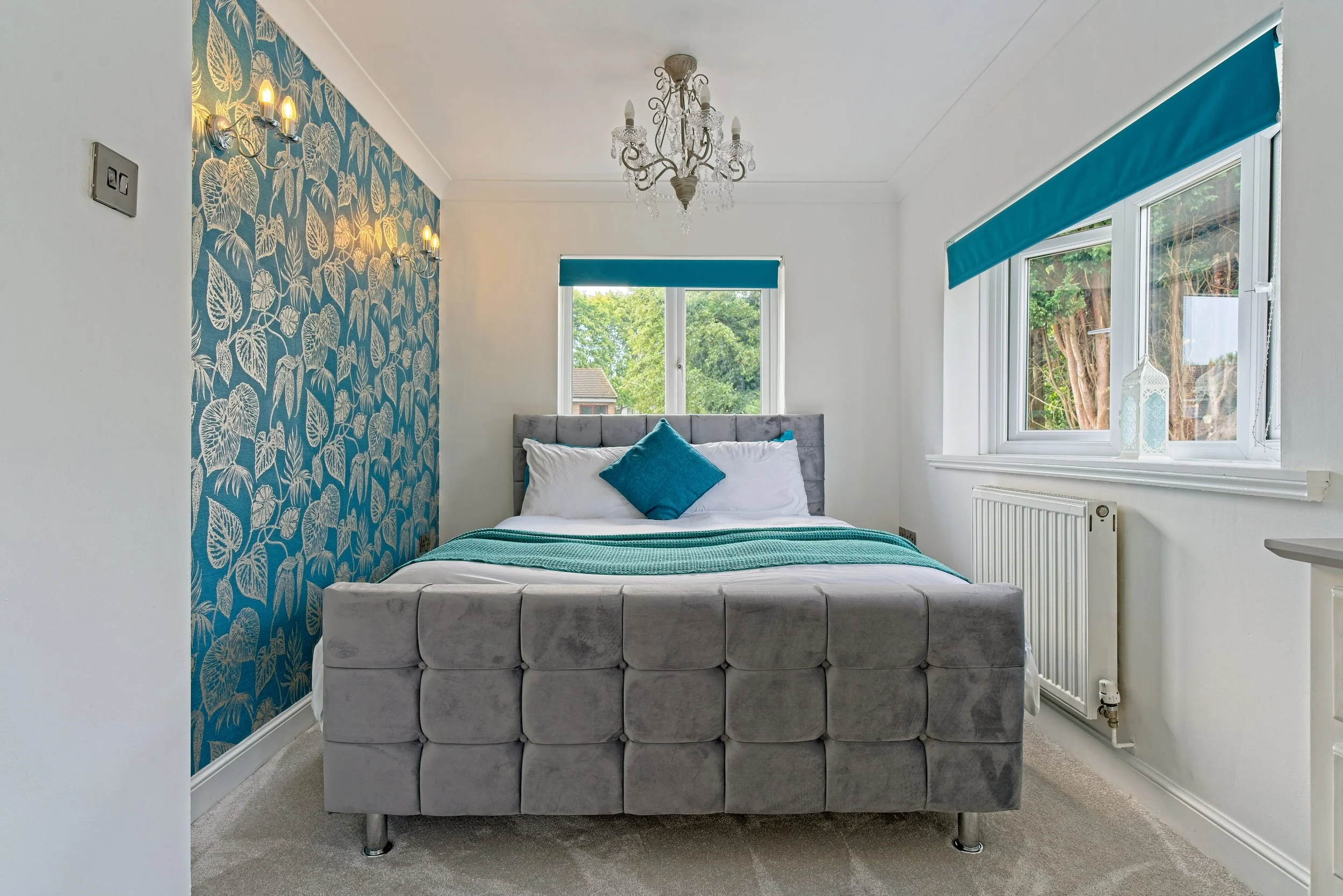 A bedroom with a gray upholstered bed, white bedding, and blue accent pillows, featuring a blue floral wallpaper wall, a chandelier, blue Roman shades on the windows, and decorative lanterns on the window sill. Property Photography Warwickshire