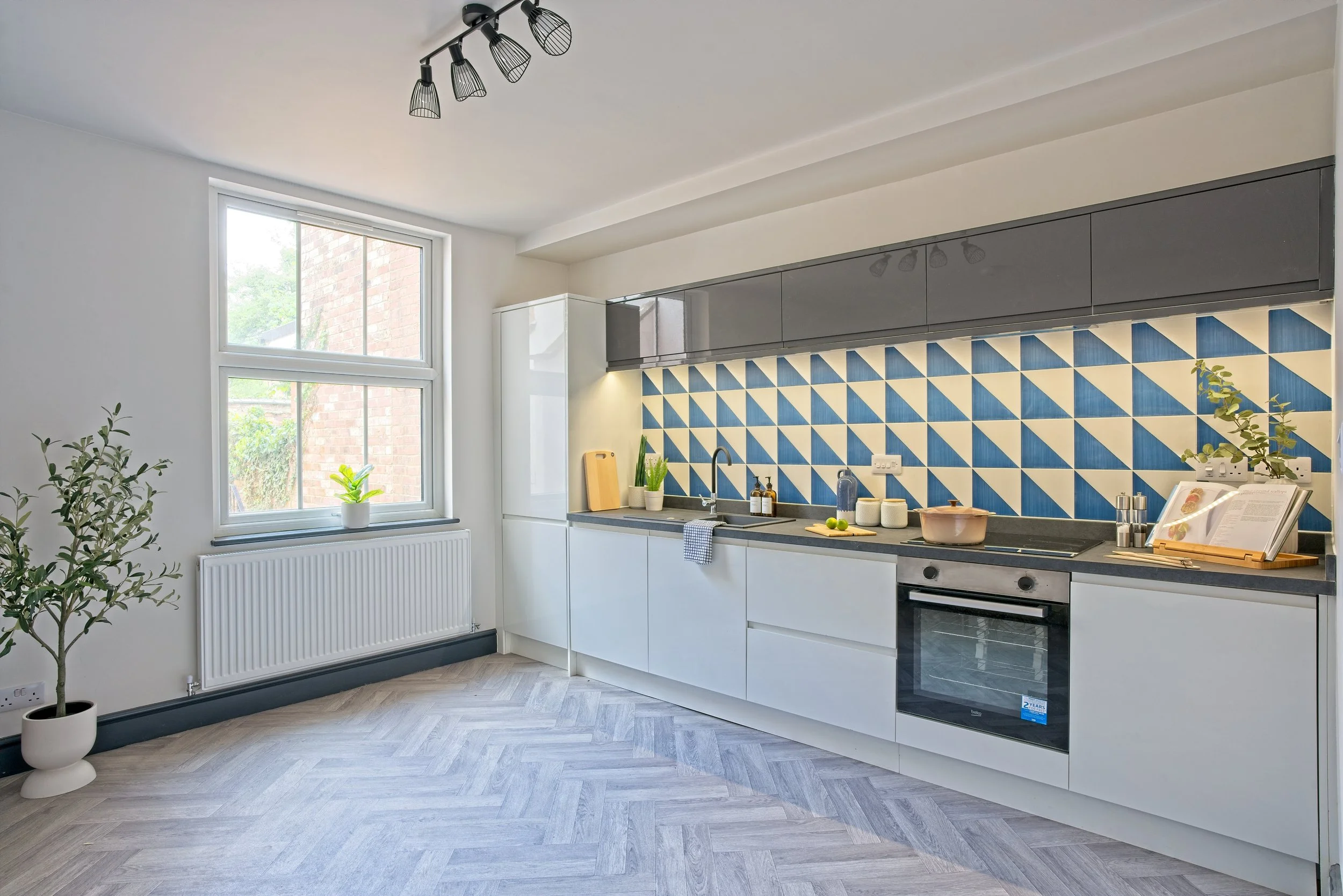 Modern kitchen with white cabinets, blue geometric tile backsplash, and gray upper cabinets, lit by ceiling spotlights and natural light from a window. Photography by photographer based in Southam, Warwickshire