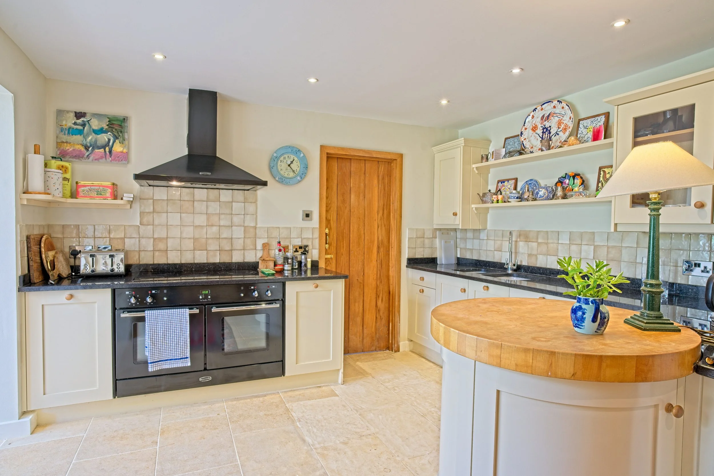 Bright kitchen with cream cabinets, black countertop, tiled backsplash, and a wooden countertop island. Appliances include a double oven and toaster. Shelves hold decorative plates and framed pictures. A blue and white vase with green plants and a gr