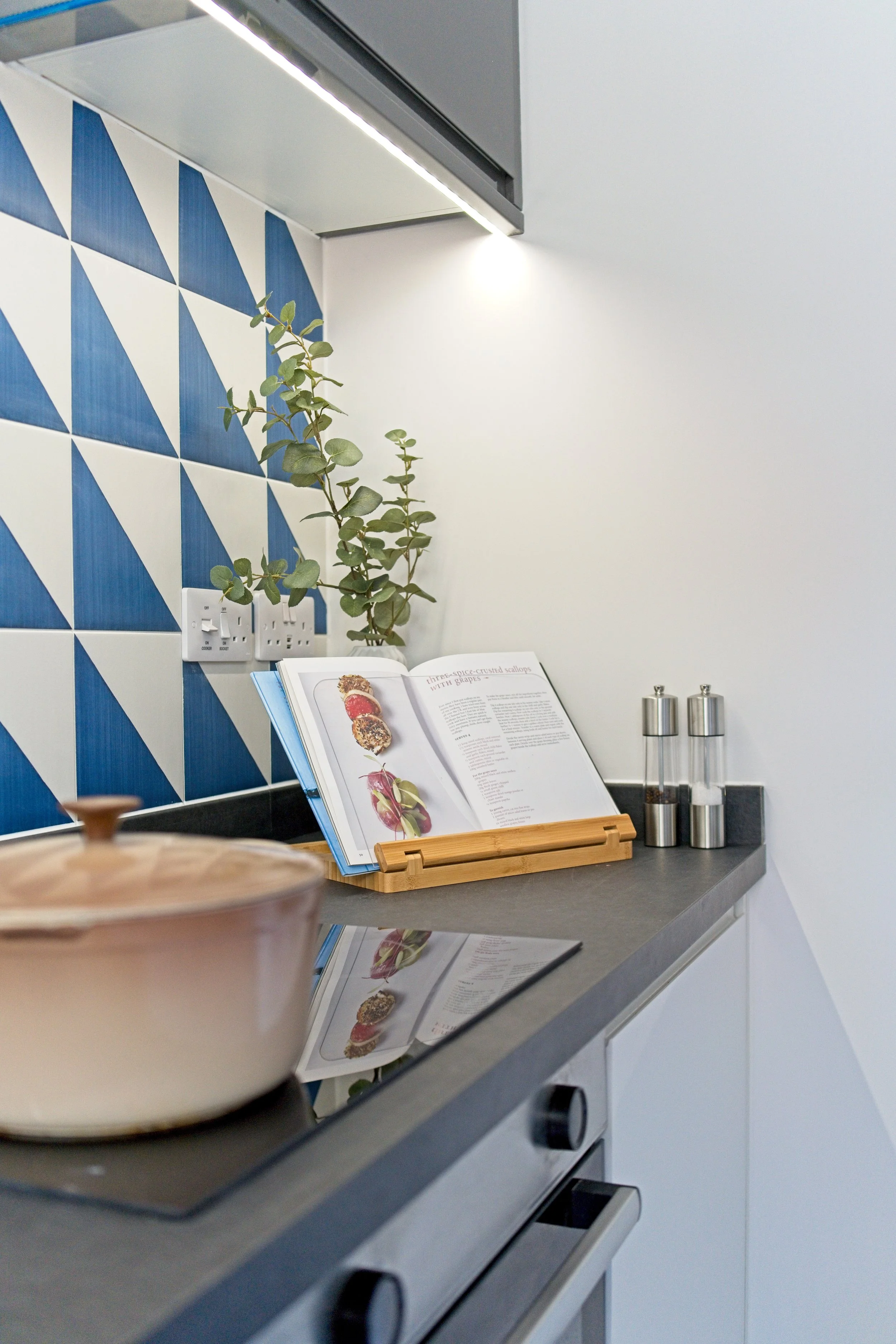 Kitchen countertop with a cookbook stand displaying an open cookbook, a potted plant, spice shakers, a pot on the stove, and a blue geometric tile backsplash. Property Photography by photographer based in Southam, Warwickshire