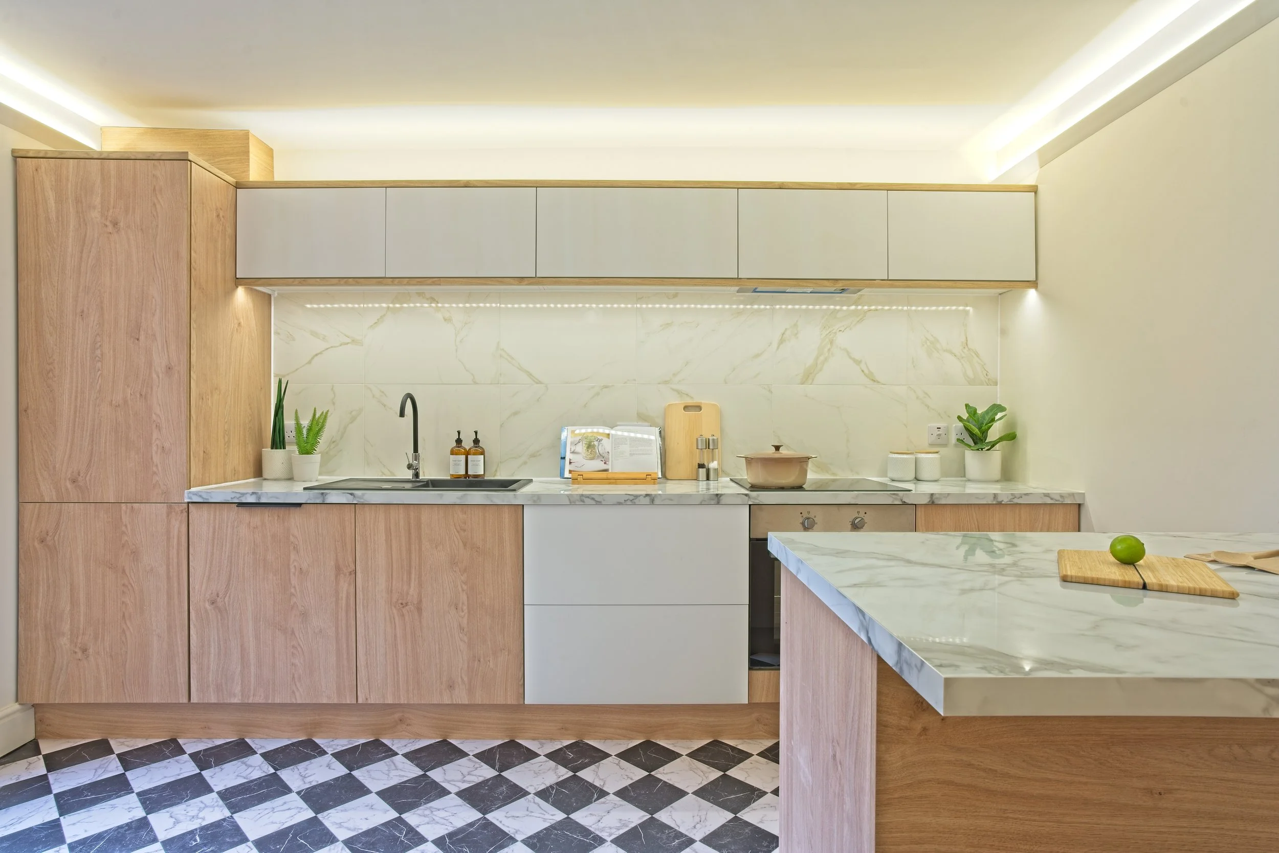 Modern kitchen with light wooden cabinets, marble countertops, and a checkered black and white floor, decorated with potted plants, a cutting board with a lime, and kitchen essentials. Photography by photographer based in Southam, Warwickshire