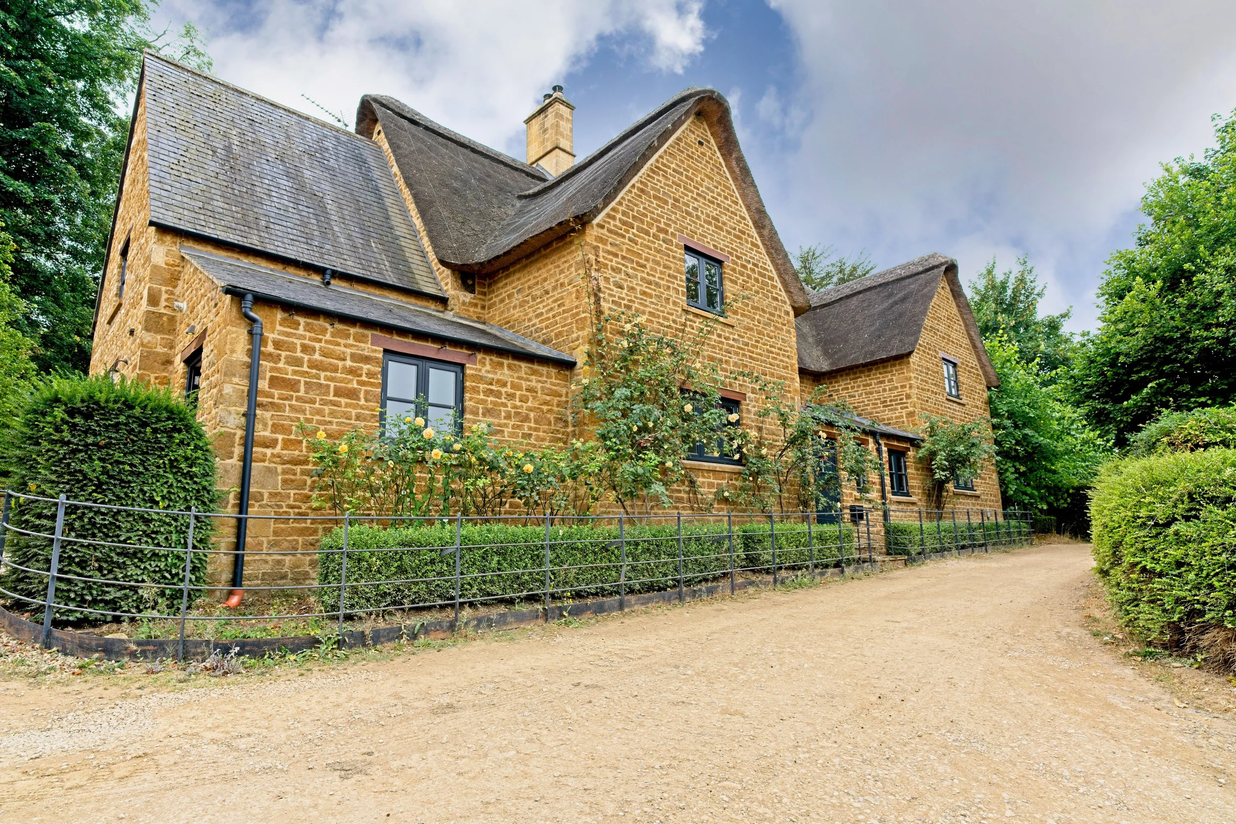 A historic stone house with thatched roof, surrounded by green bushes and trees, with a dirt driveway in front and a cloudy sky overhead. Property photography by photographer based in Southam, Warwickshire