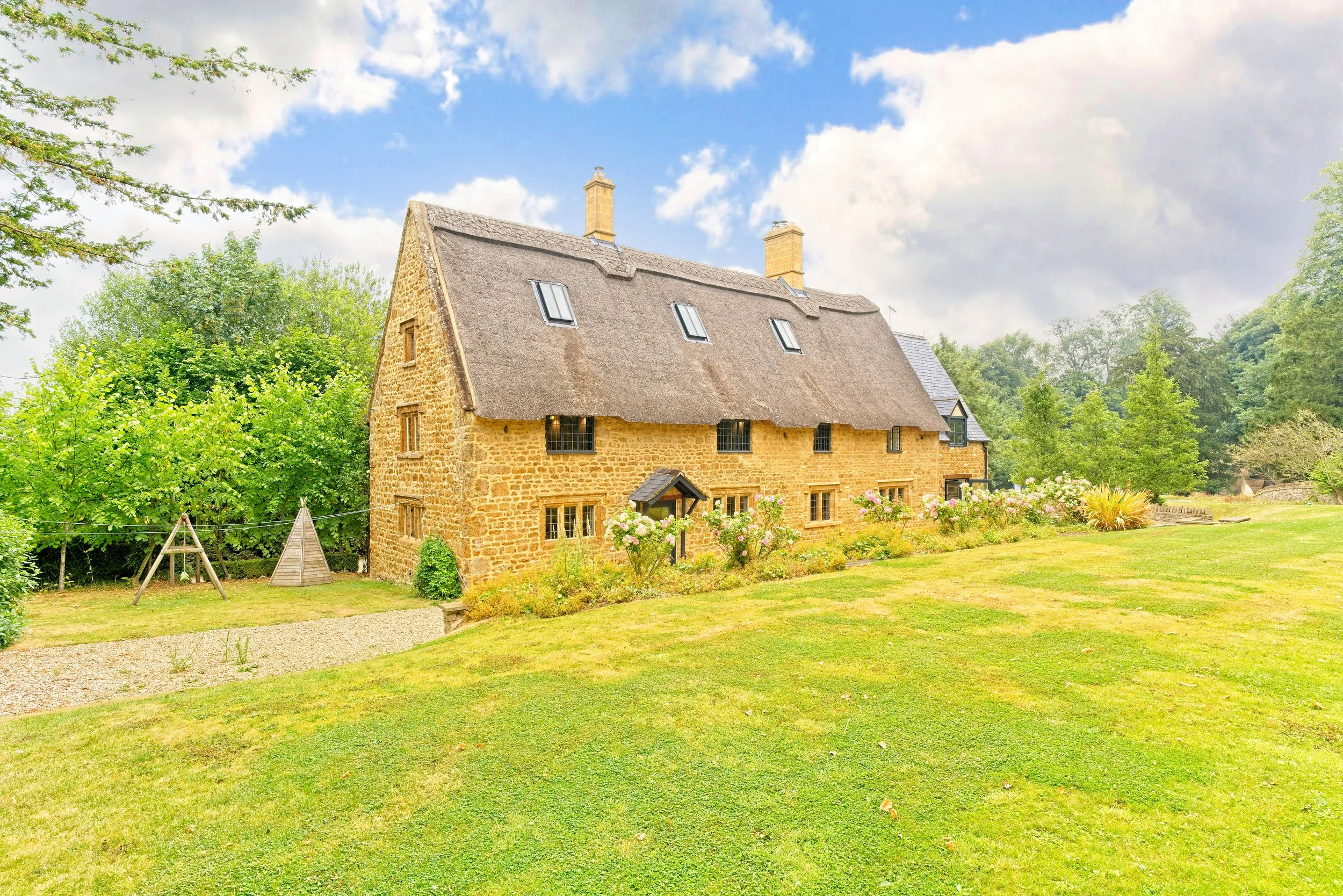 Stone cottage with a thatched roof surrounded by a green lawn and trees, under a partly cloudy sky. Property Photography by photographer based in Southam, Warwickshire