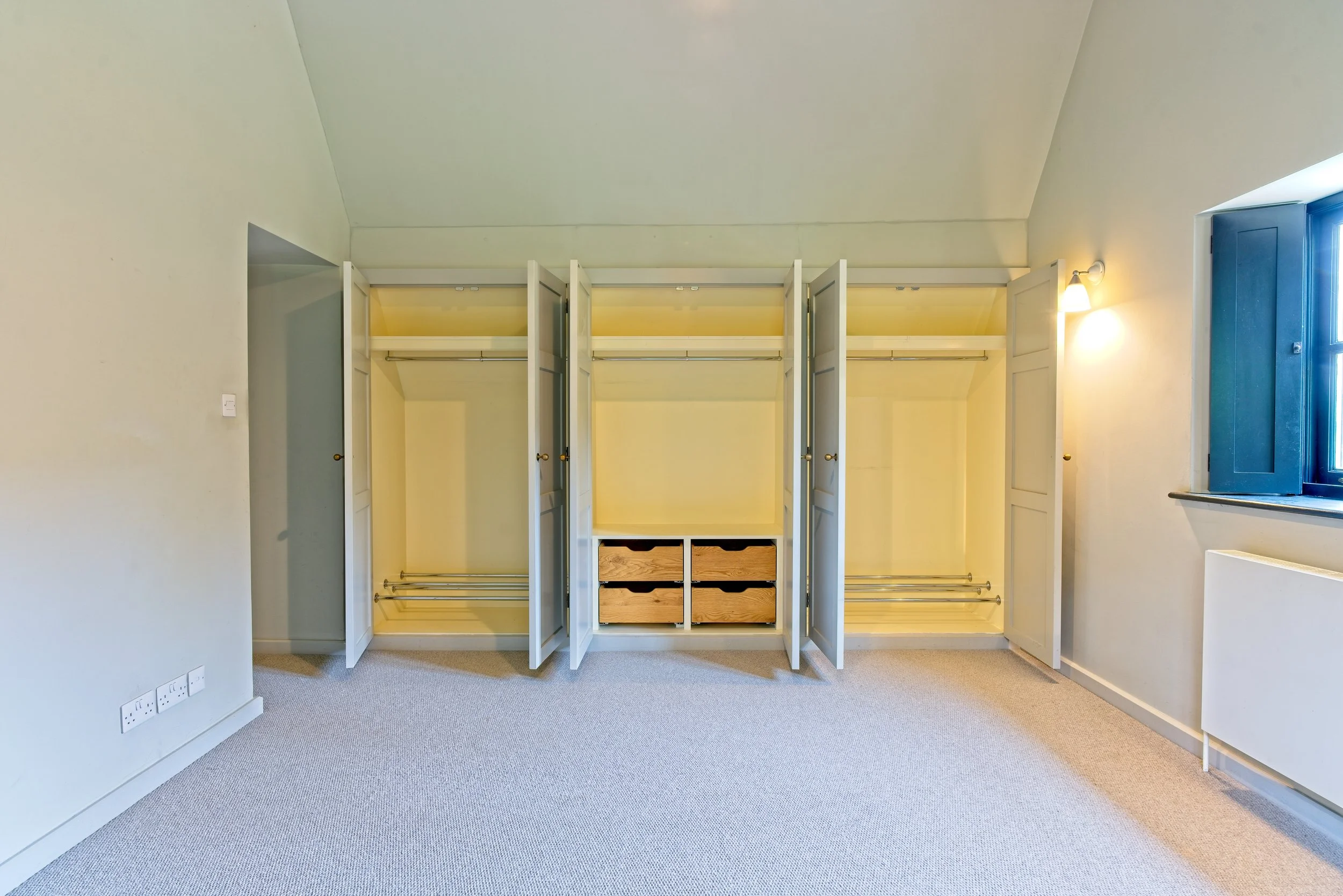 Empty built-in wardrobe with open doors, wooden drawers in the middle, and a window with blue shutters in a room with beige walls and carpeted floor. Property photography by photographer based in Southam, Warwickshire